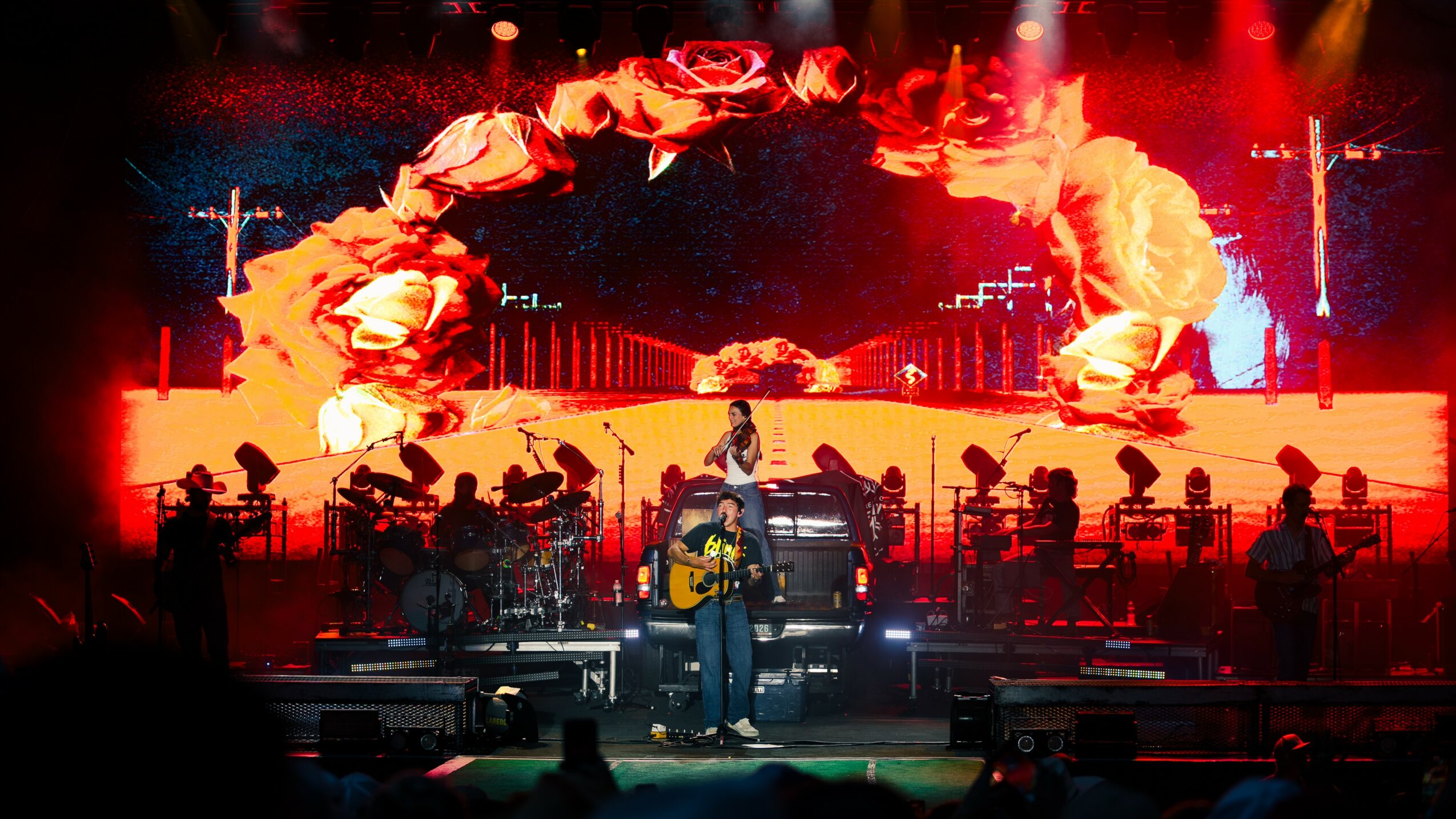 Musician performing on stage with a large digital screen displaying a floral arch and flames behind, with multiple instruments and musicians.