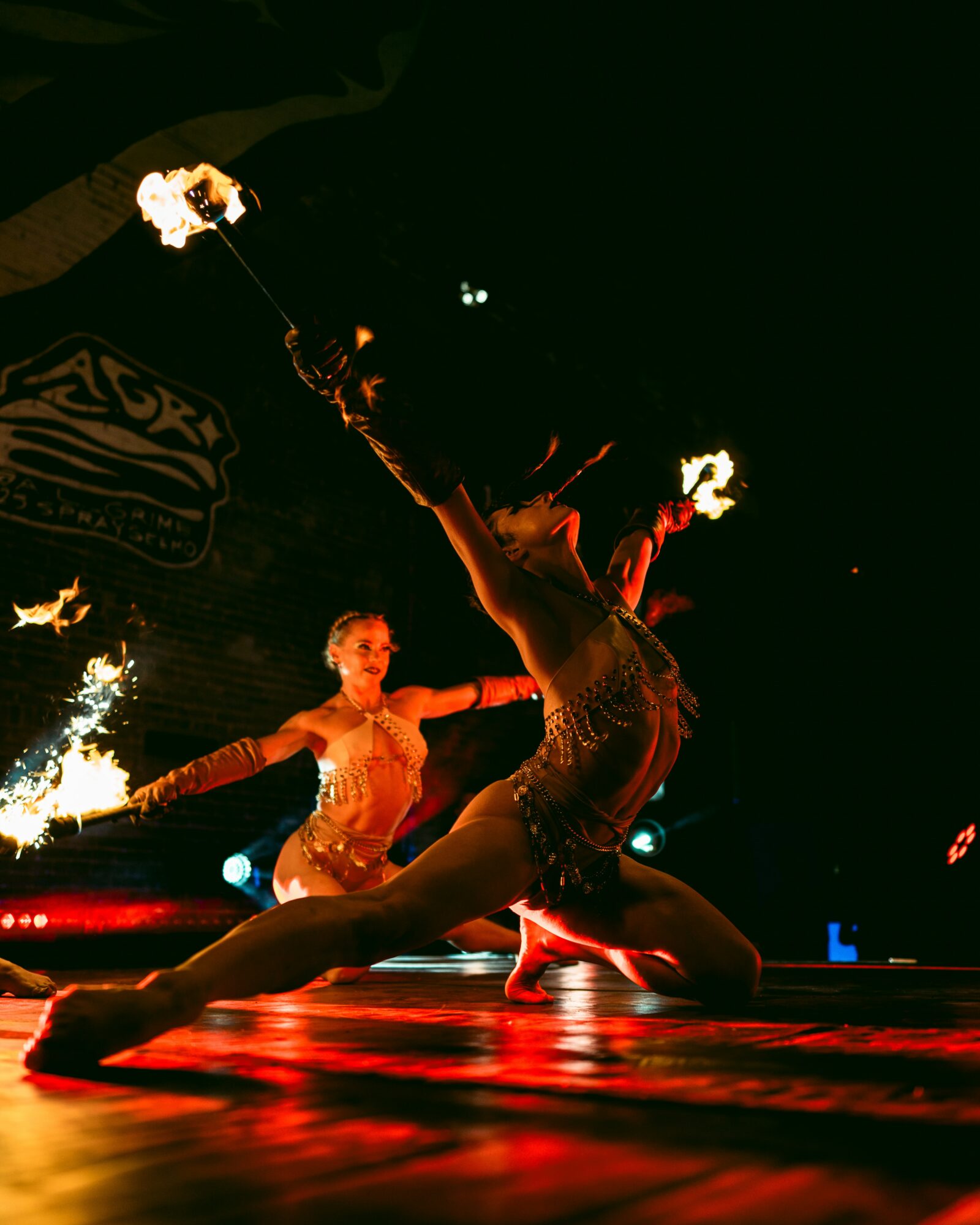 Two performers with fire props on stage, one in a split position, illuminated by stage lights, dark background.