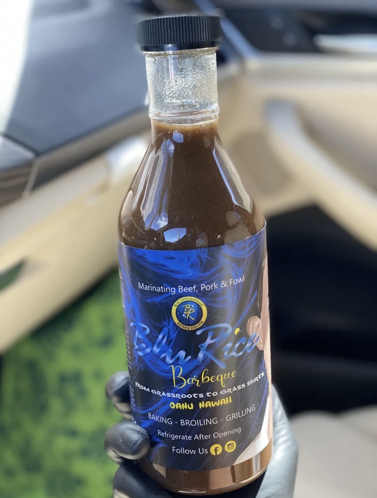 Bottle of barbecue sauce inside a car, with a blue label and black cap, on a beige car interior seat.