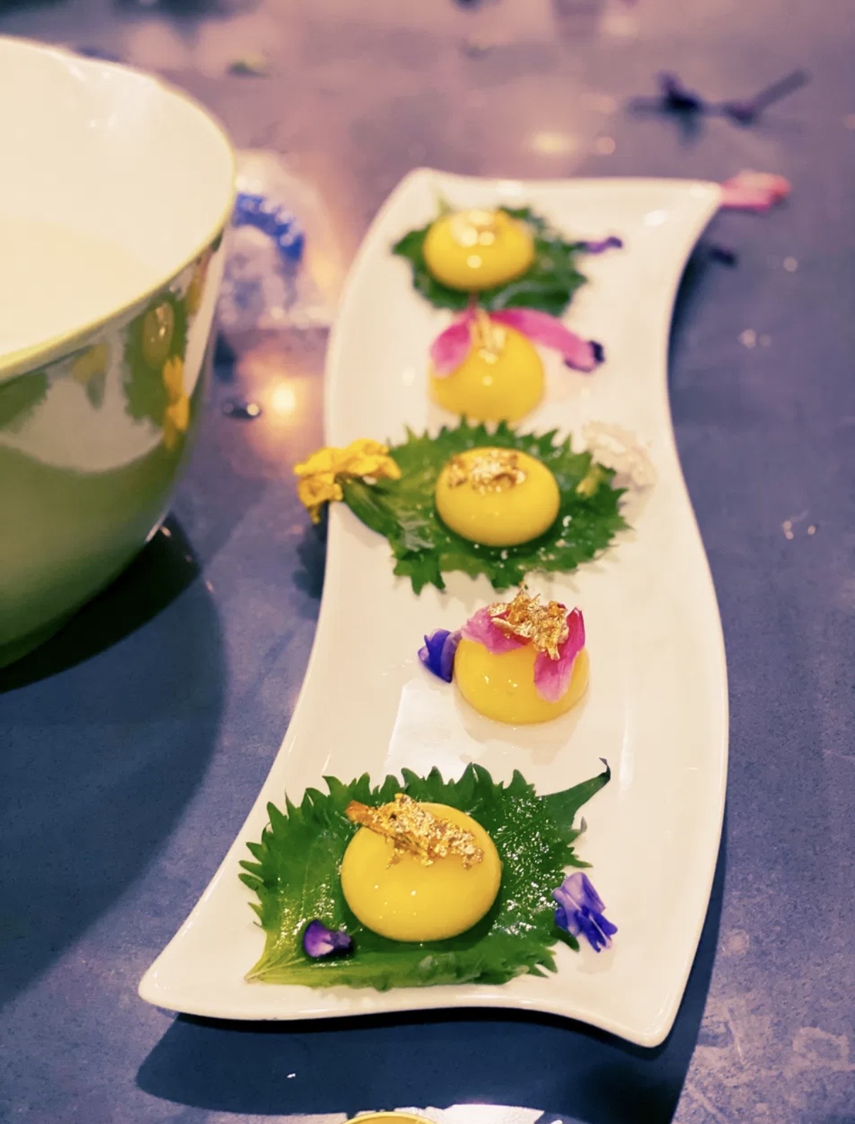 Yellow desserts with green leaves and colorful flower decorations on a white curved plate, with a bowl nearby.
