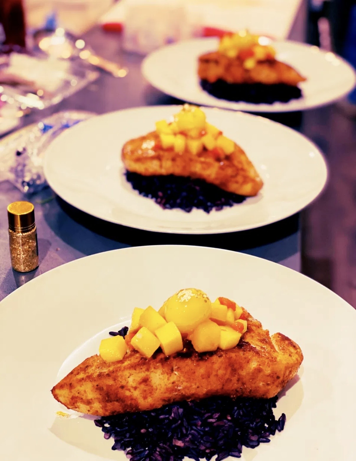 Three plates with slices of pie topped with fruit and mango pieces, on a table with utensils and a spice container.