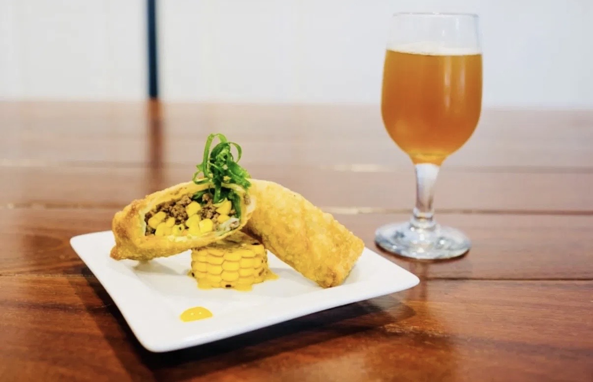 Fried fish fillet with garnish, yellow corn, and a glass of beer on a wooden table.