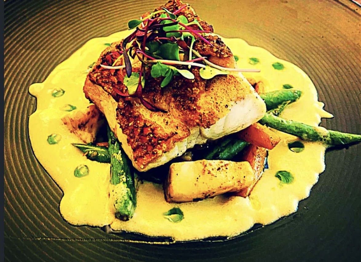 Pan-seared fish fillet on green beans, potatoes, and creamy sauce, garnished with microgreens.