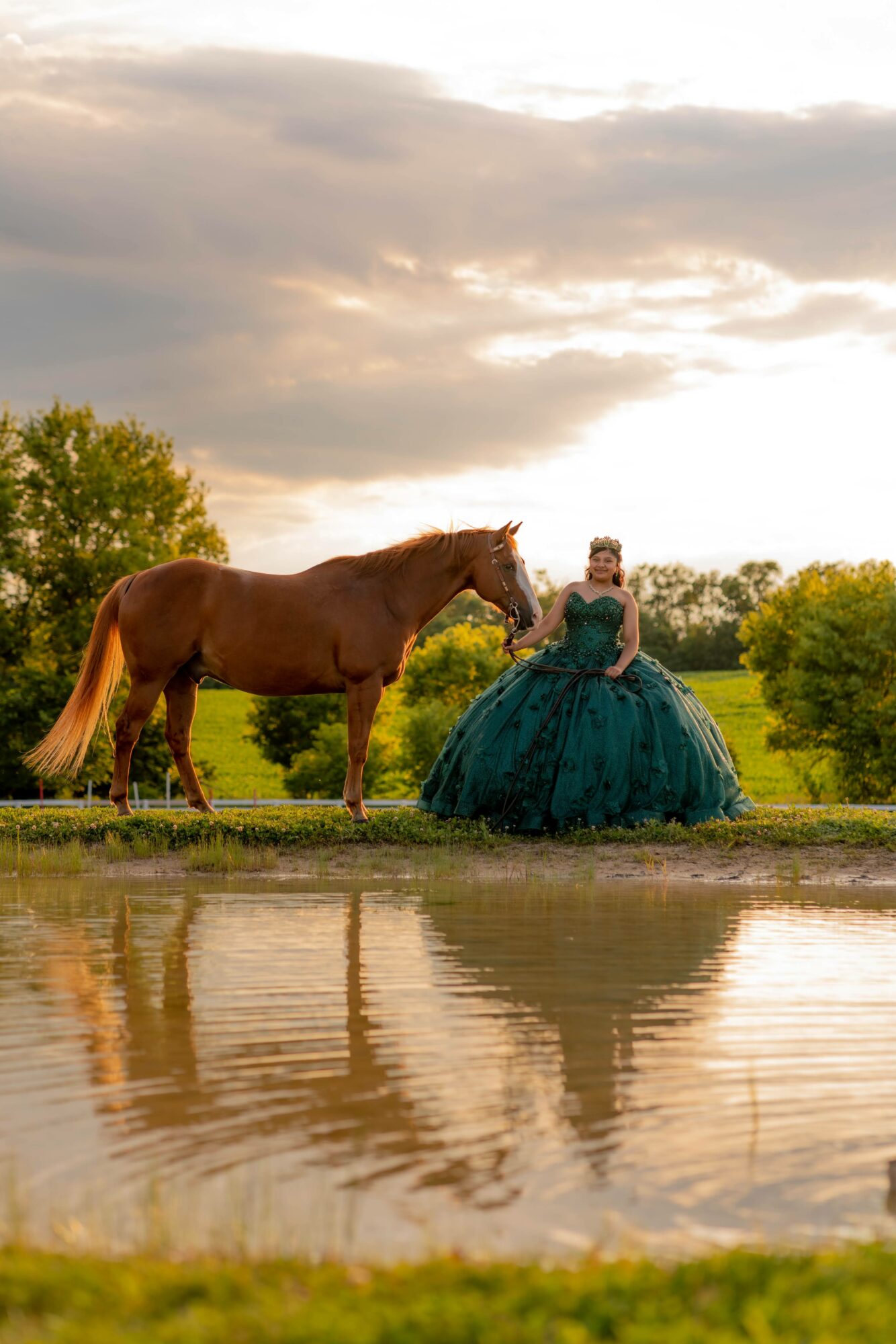 A woman in a teal ball gown stands near a brown horse by a pond with trees and cloudy sky in the background.