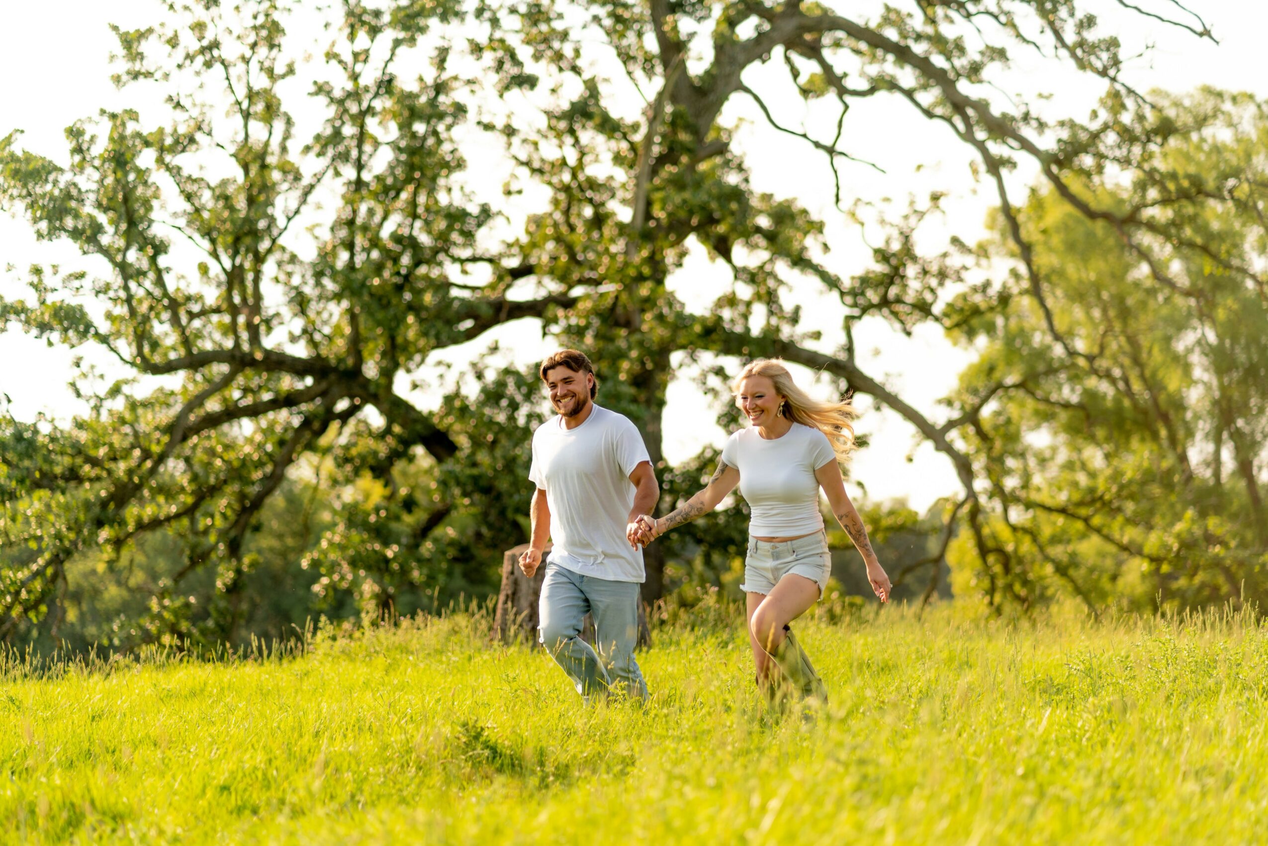 A couple walking hand in hand in a grassy field with trees in the background, smiling and enjoying the outdoors.