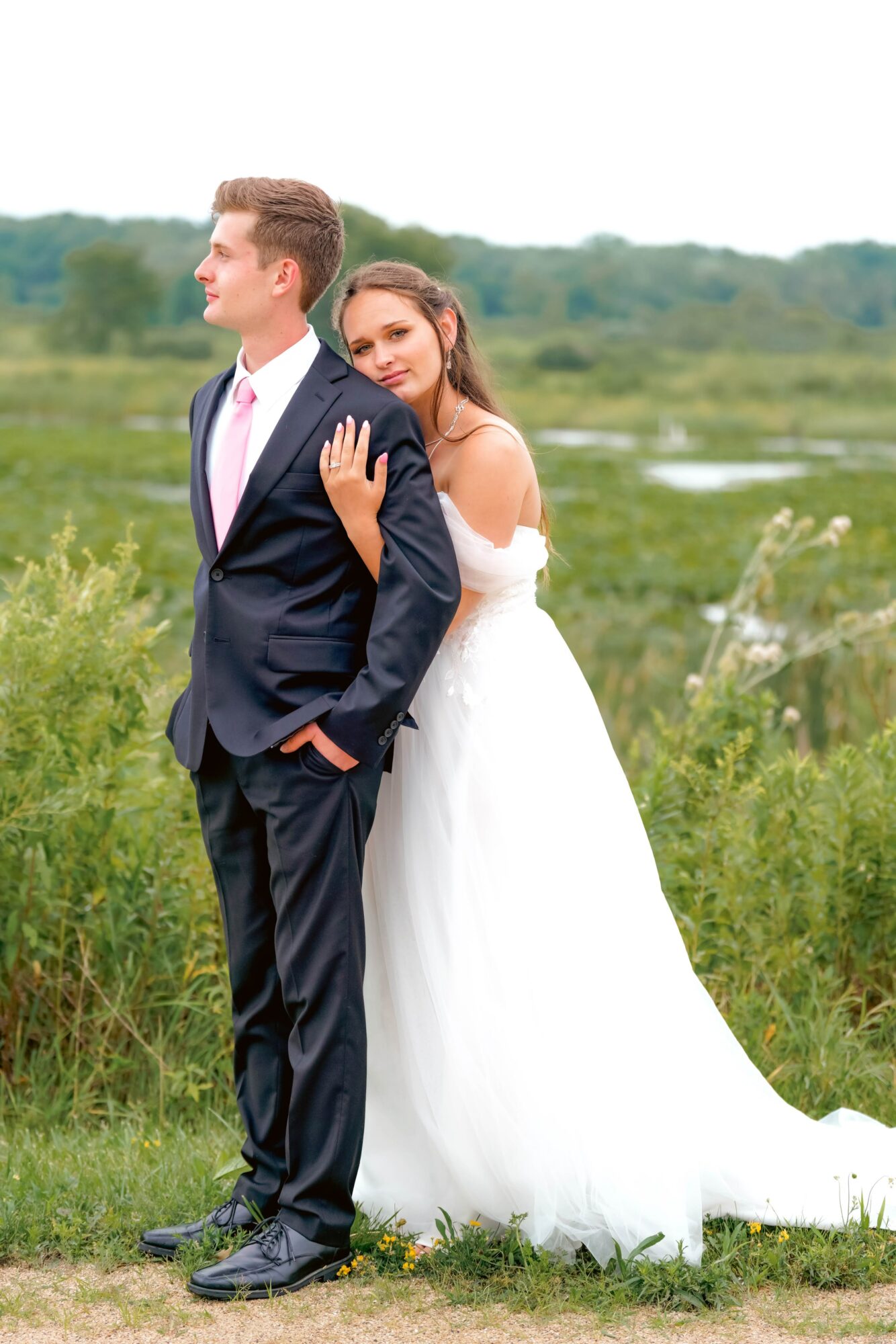 Bride and groom outdoors, bride leaning on groom's shoulder, green landscape background, wedding attire, calm expressions.