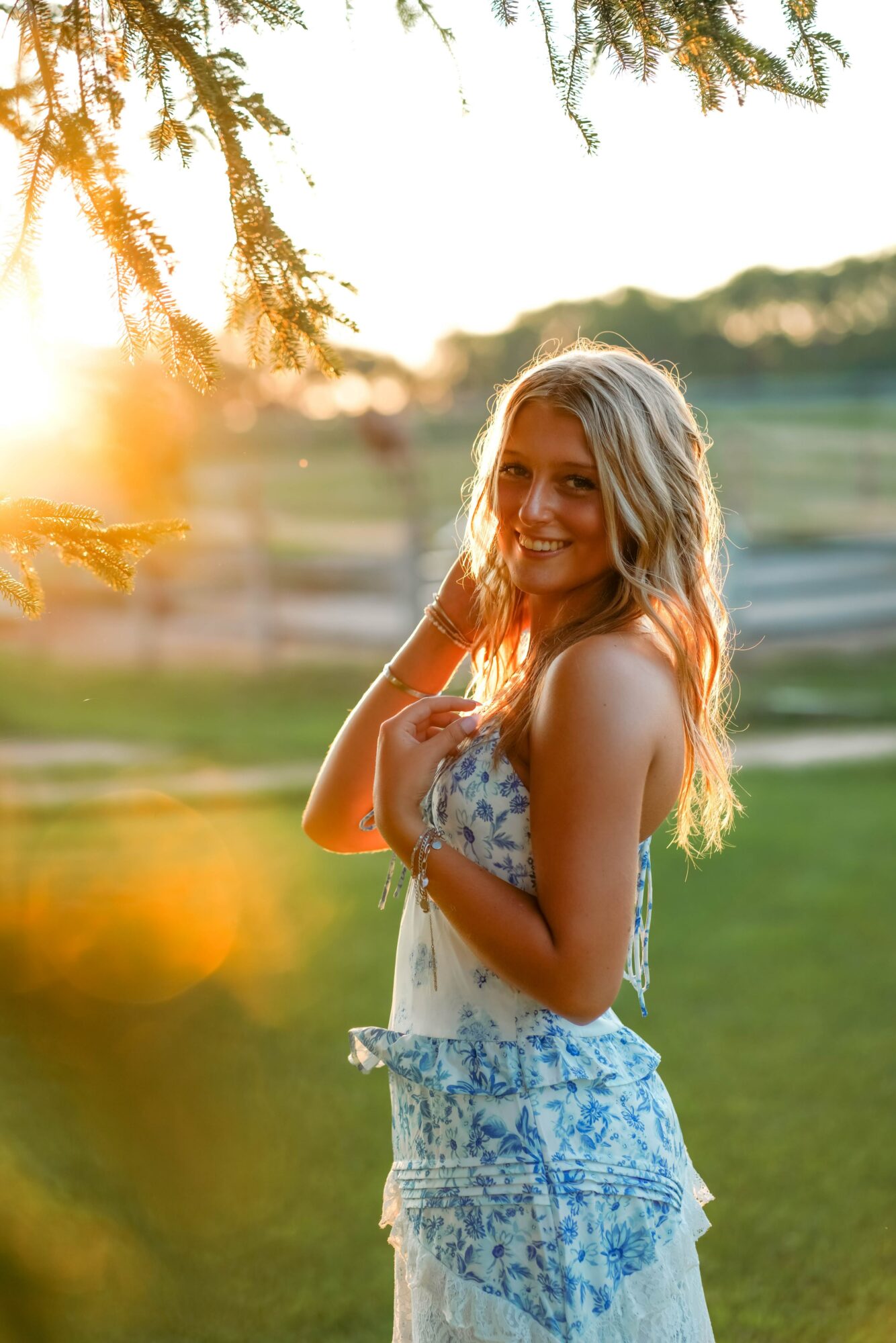 Smiling woman with long hair in a floral dress outdoors during sunset, with trees and grass in background.