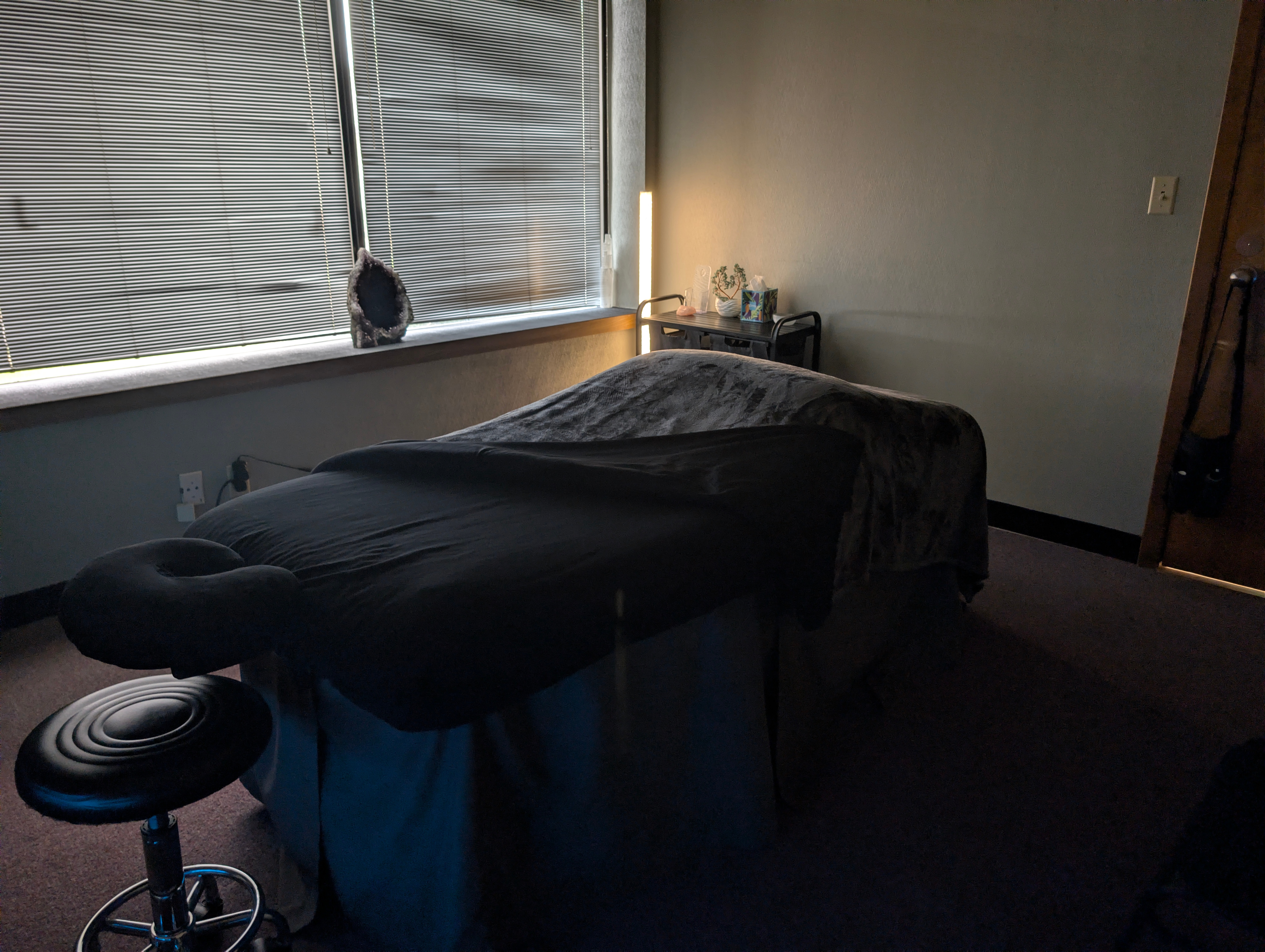 Massage table in a dimly lit room with a window, a small table, and a stool.