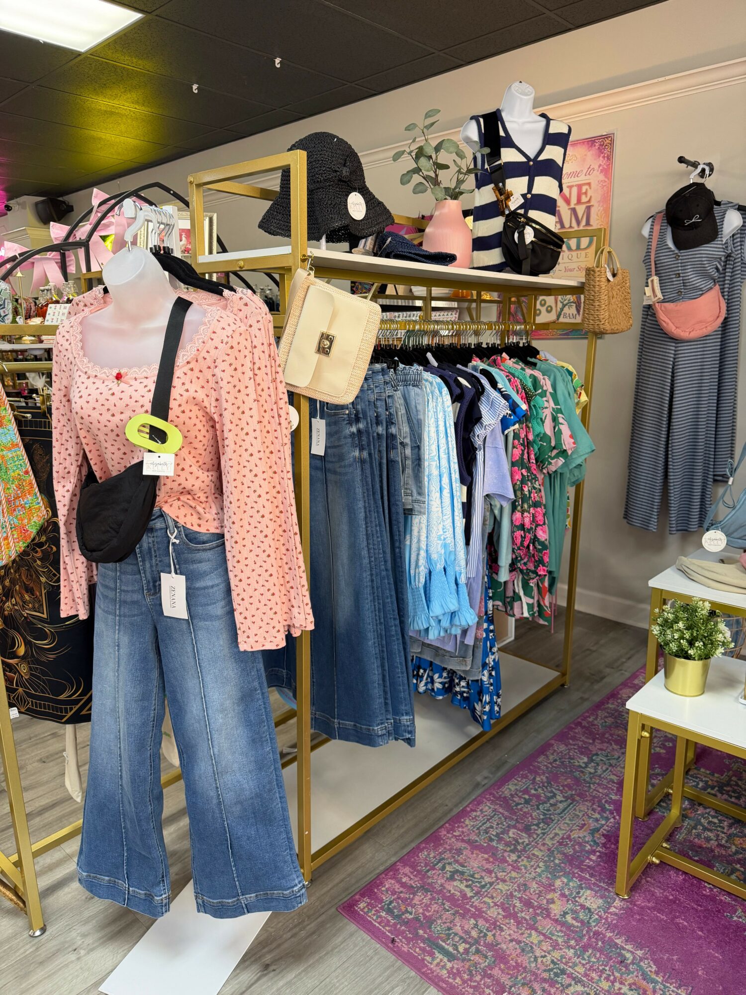 Clothing display with jeans, shirts, and dresses on a gold rack in a boutique, with a pink rug and other clothing racks visible.