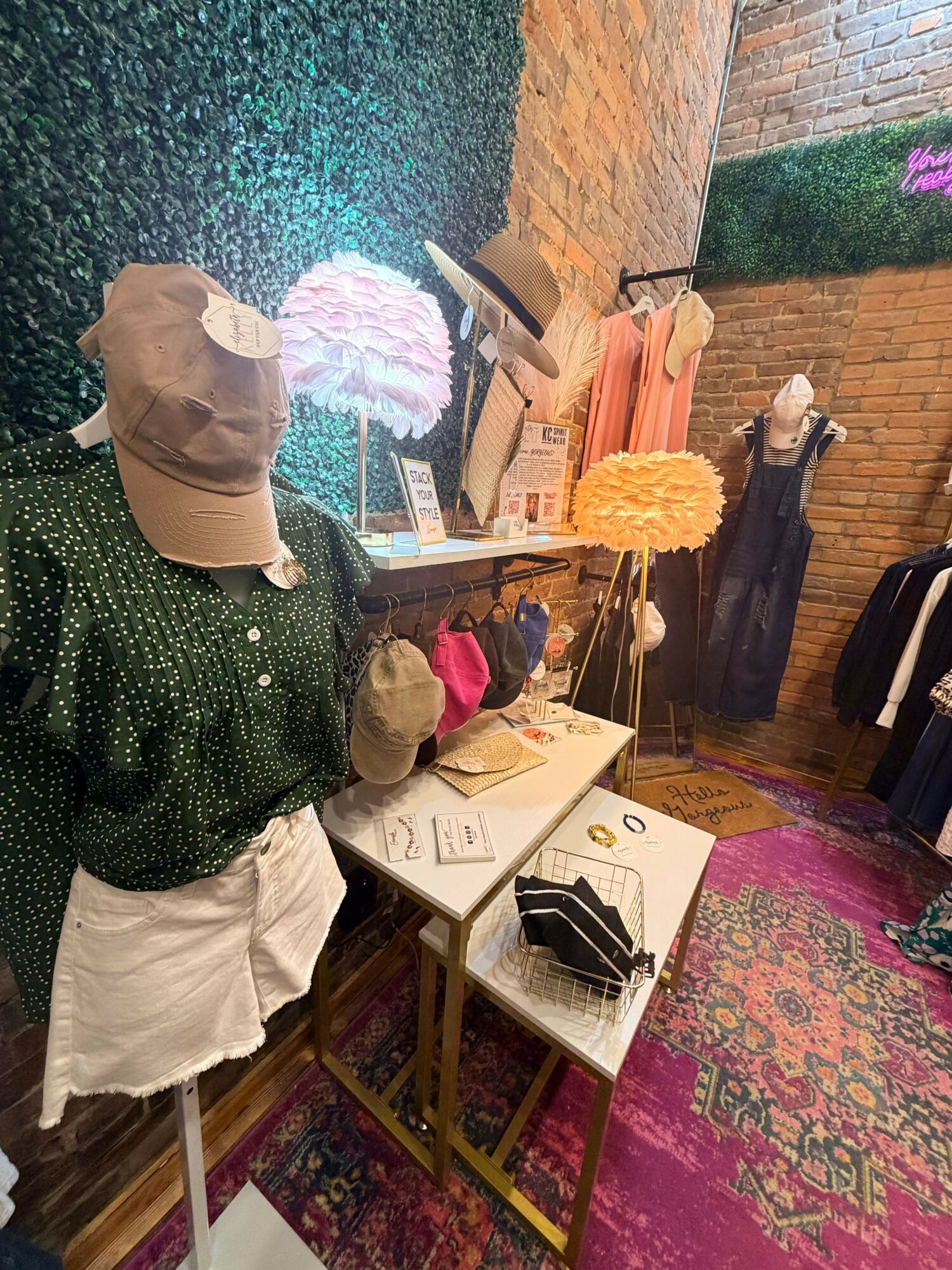 Person wearing a beige hat and green jacket browsing clothing at an indoor market or store, with tables and racks of clothes nearby.