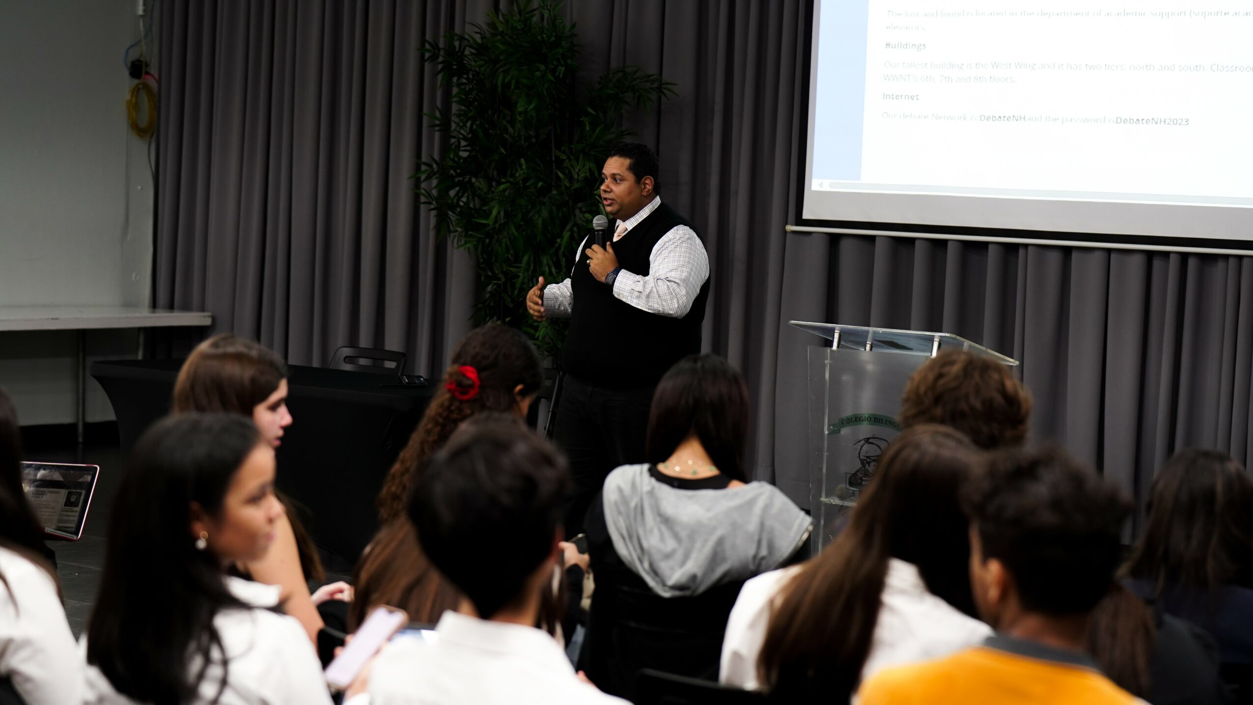 Man speaking to an audience in a conference room with a presentation screen, audience members listening attentively.