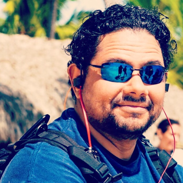 Man with curly hair, sunglasses, and a backpack outdoors smiling, with trees and rocks in background.