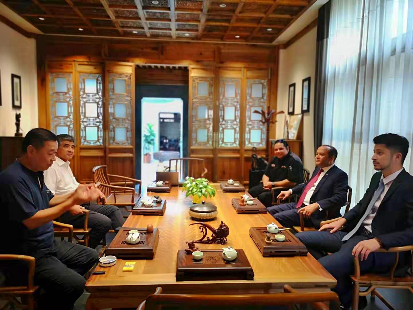 Six people seated around a wooden conference table in a room with wooden walls and large windows, engaged in discussion.
