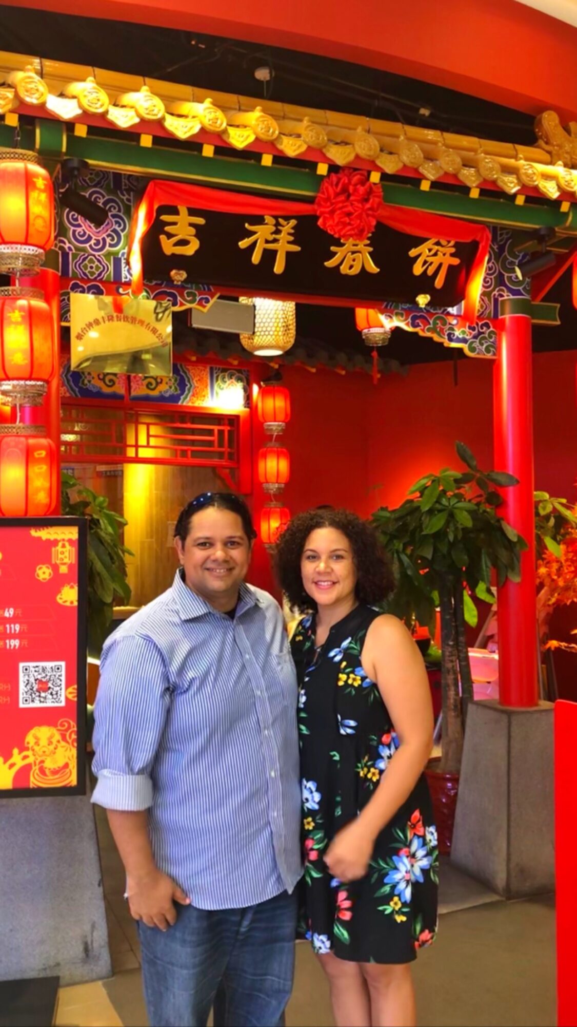 Two people standing in front of a decorated Chinese-themed entrance with red lanterns and plants.