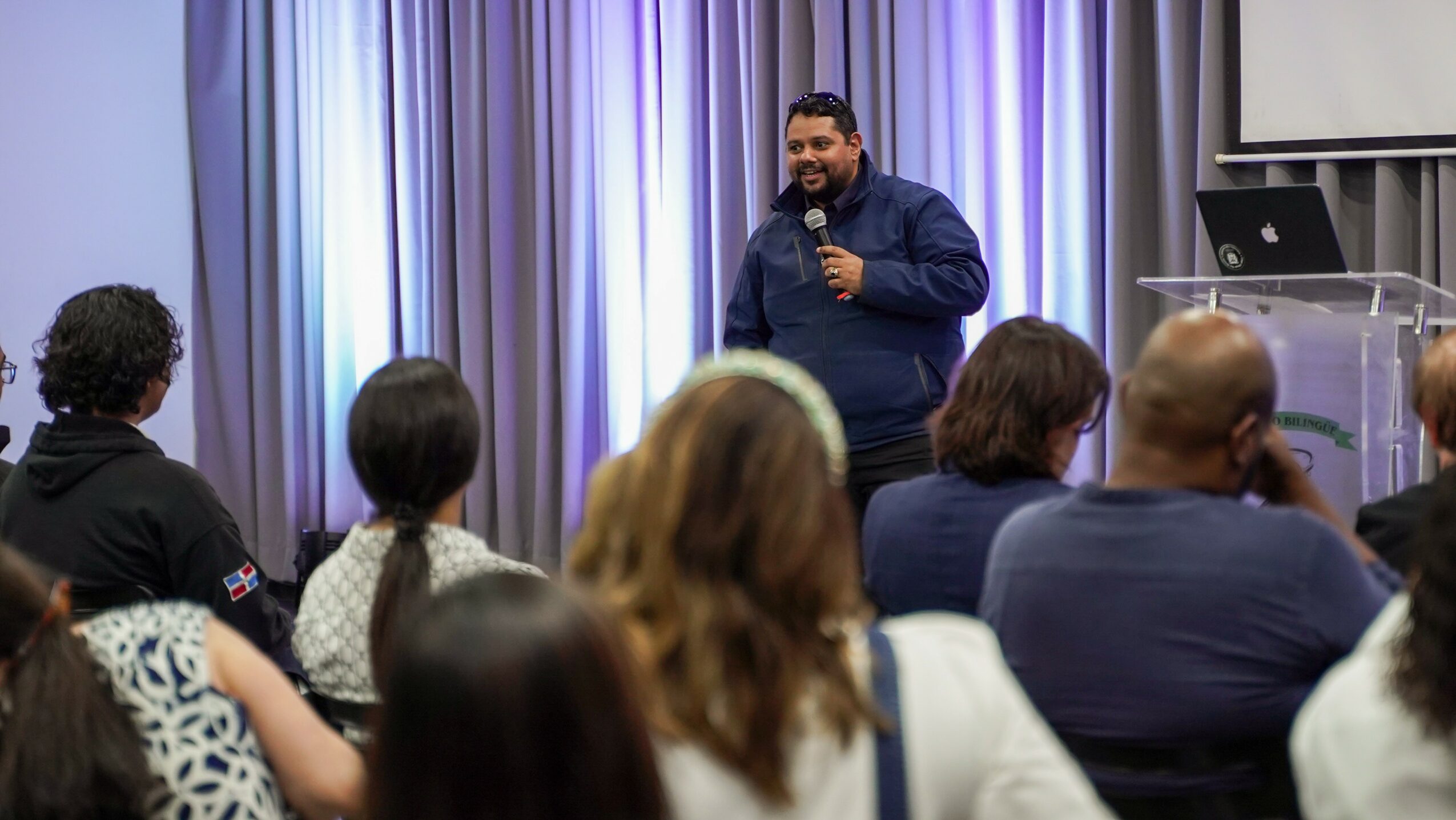 Person speaking into microphone in front of seated audience in a room with curtains and a podium.