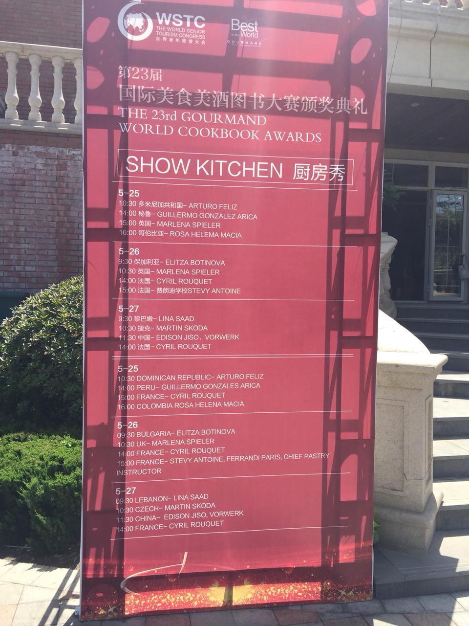 Pink signboard listing food and event details, with shadows cast on it, outdoors near a building and stairs.