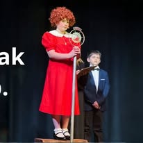 A woman in a red dress and a child in a suit stand on stage with a dark background.