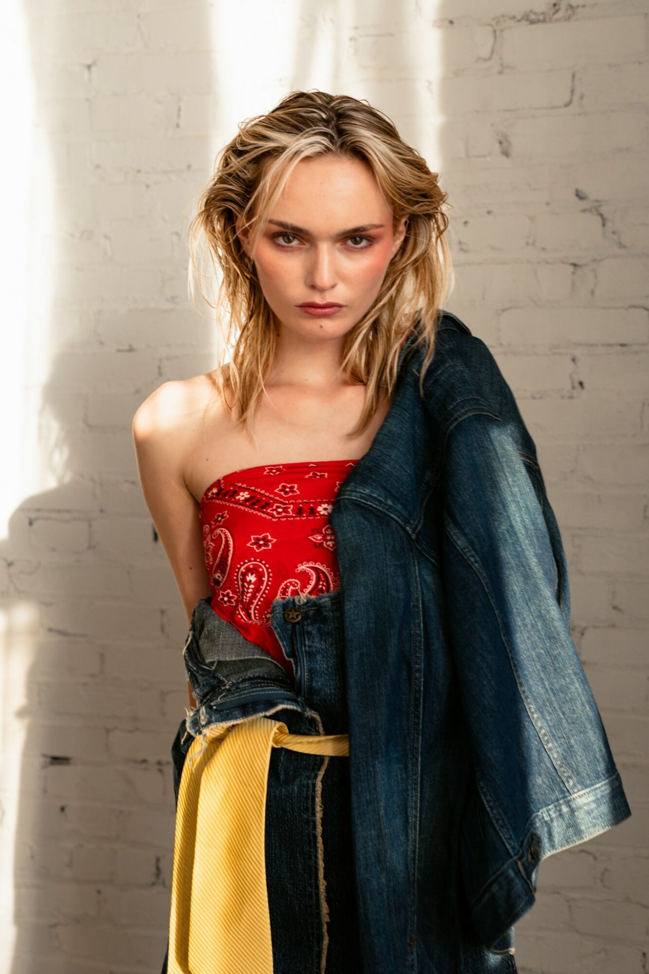 Young woman with blonde hair wearing a red top and denim jacket, standing against a white brick wall.