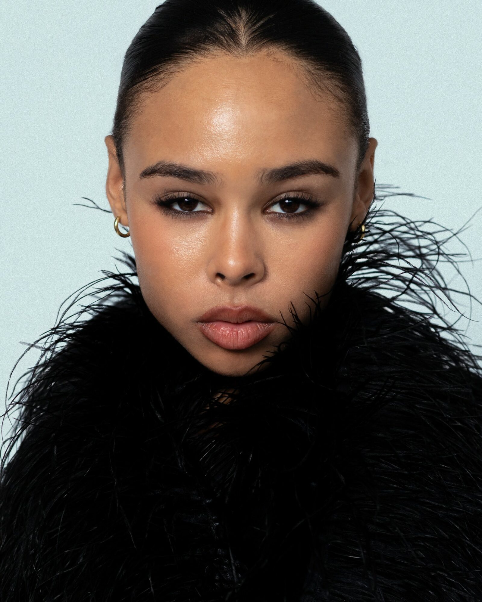 Close-up of a woman with slicked-back hair, wearing a black feathered garment, against a light background.