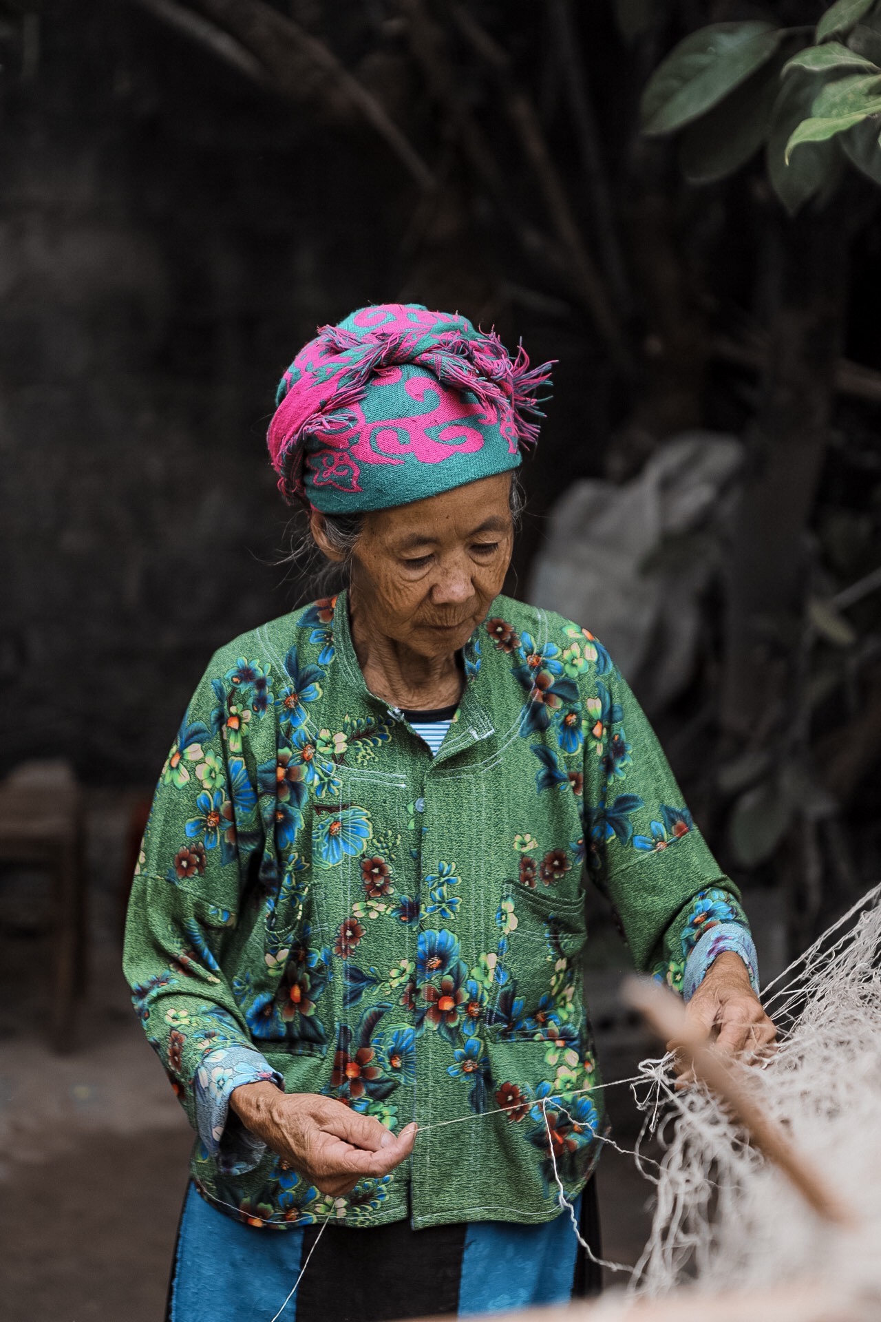 Elderly woman wearing a colorful headscarf and patterned clothing, looking down at her hands outdoors.