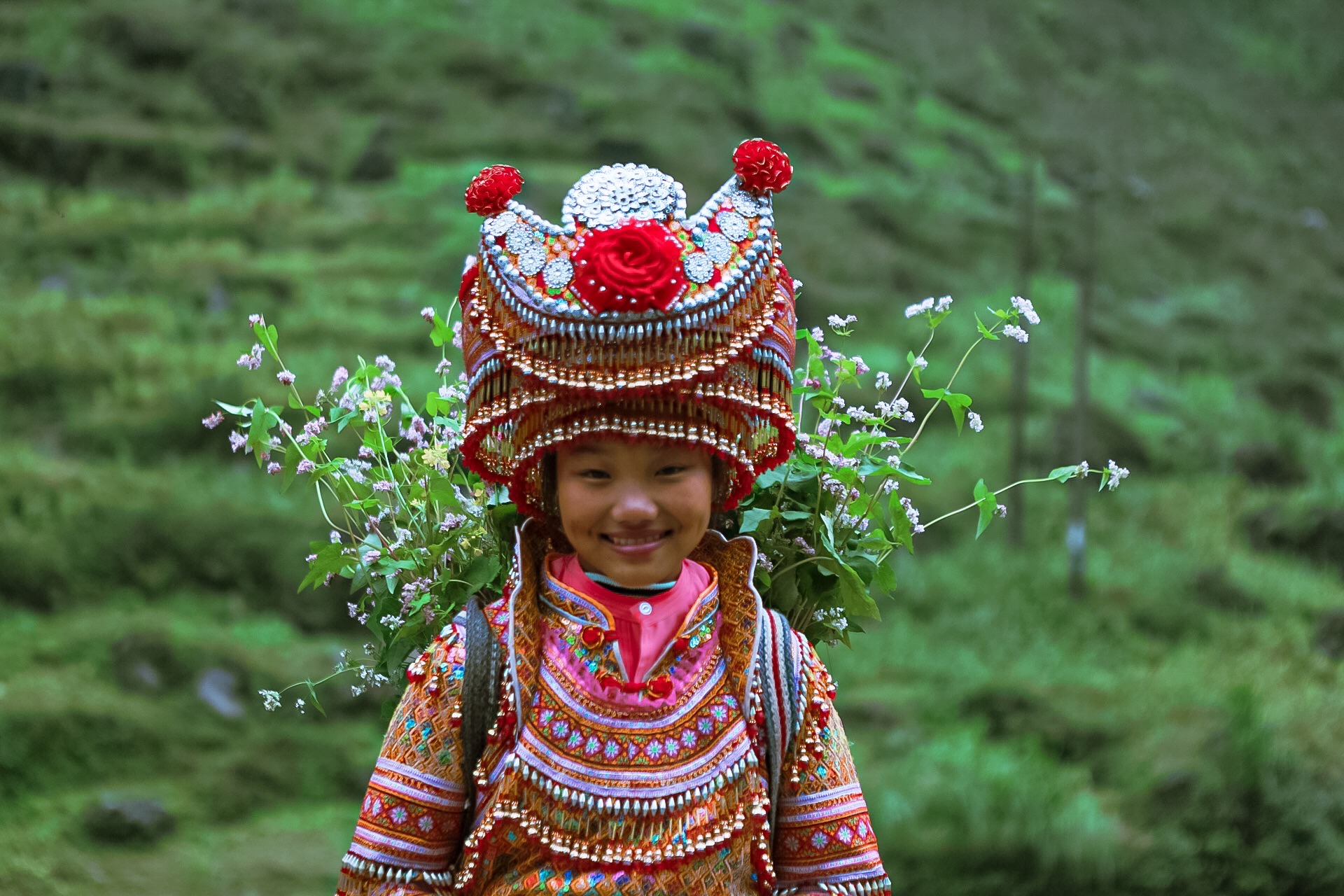 Child wearing a colorful traditional costume and elaborate headdress, standing outdoors with green landscape background.