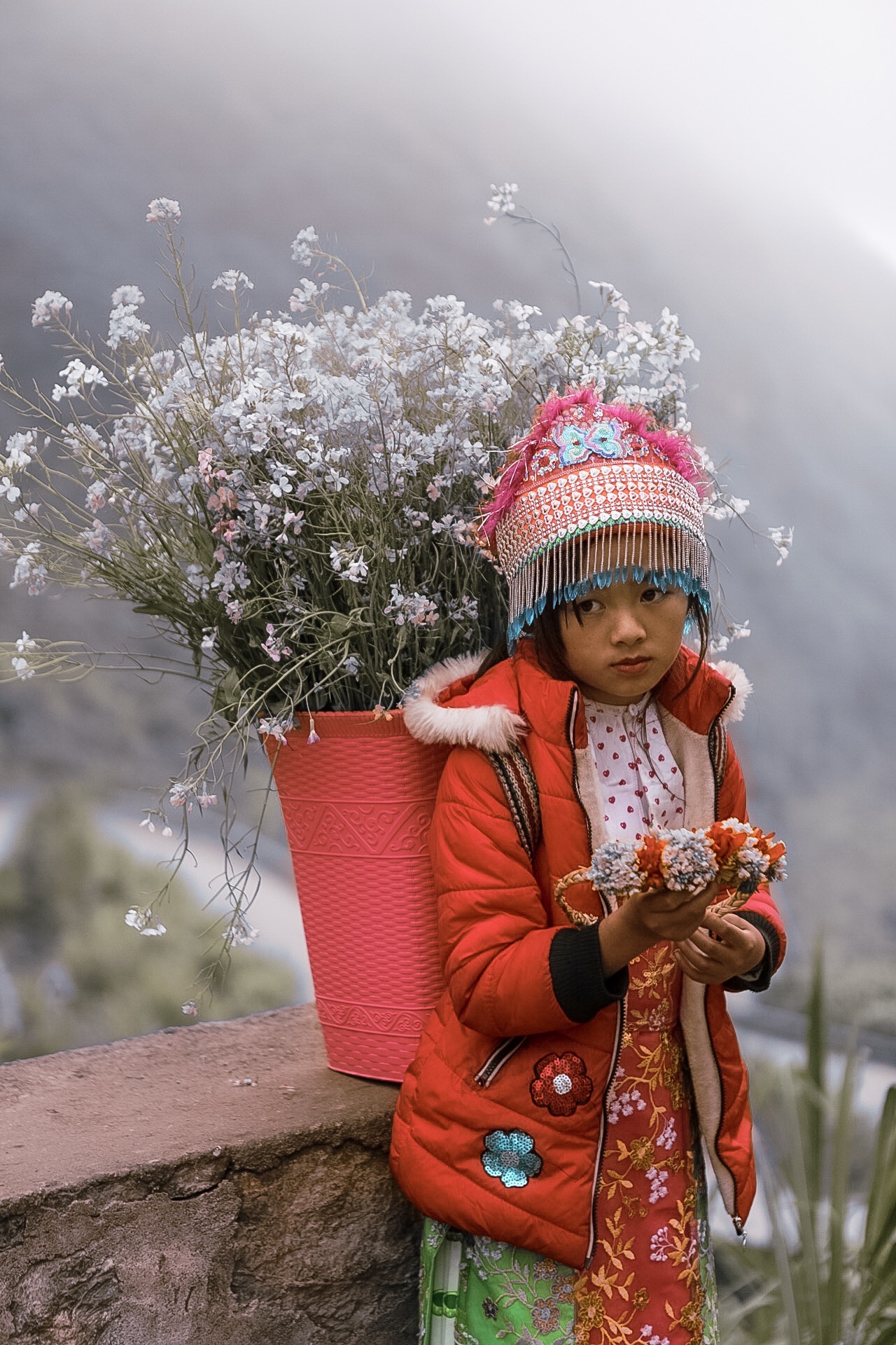 Child in red coat and colorful hat holding flowers, with a large pink basket of white flowers behind them, outdoors.