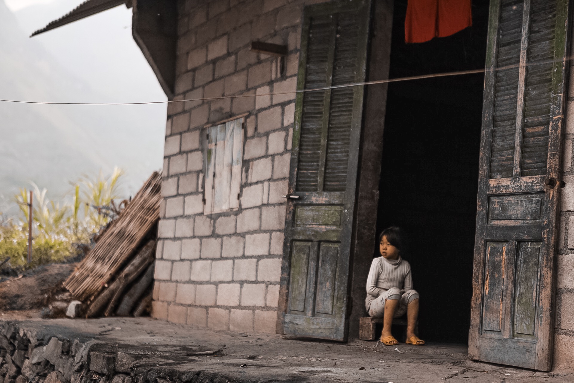 Child sitting in doorway of a brick house with open wooden shutters, laundry hanging above, and a rural background.