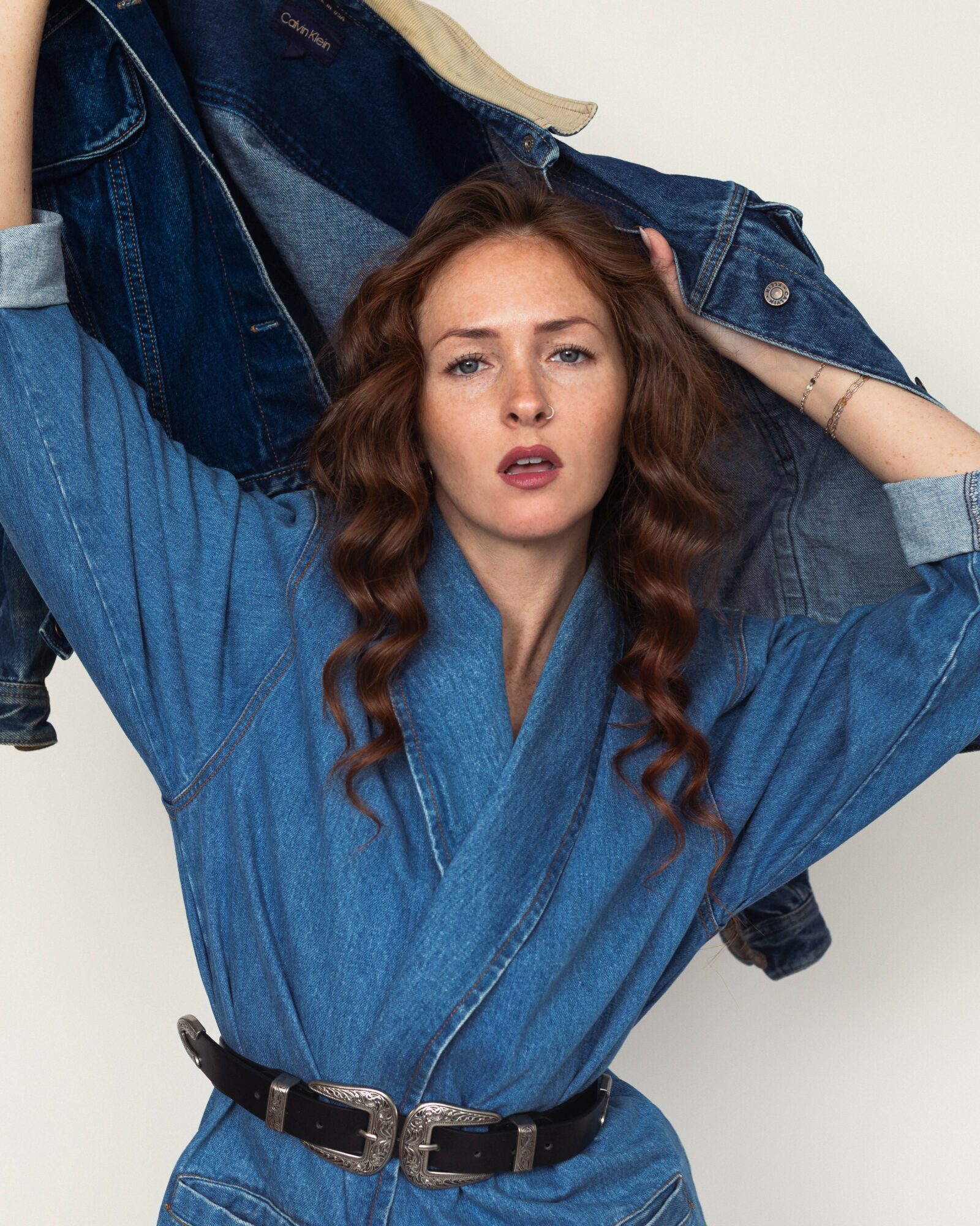 Woman with long wavy hair wearing a denim jacket, holding a jacket on her shoulder, looking at the camera.