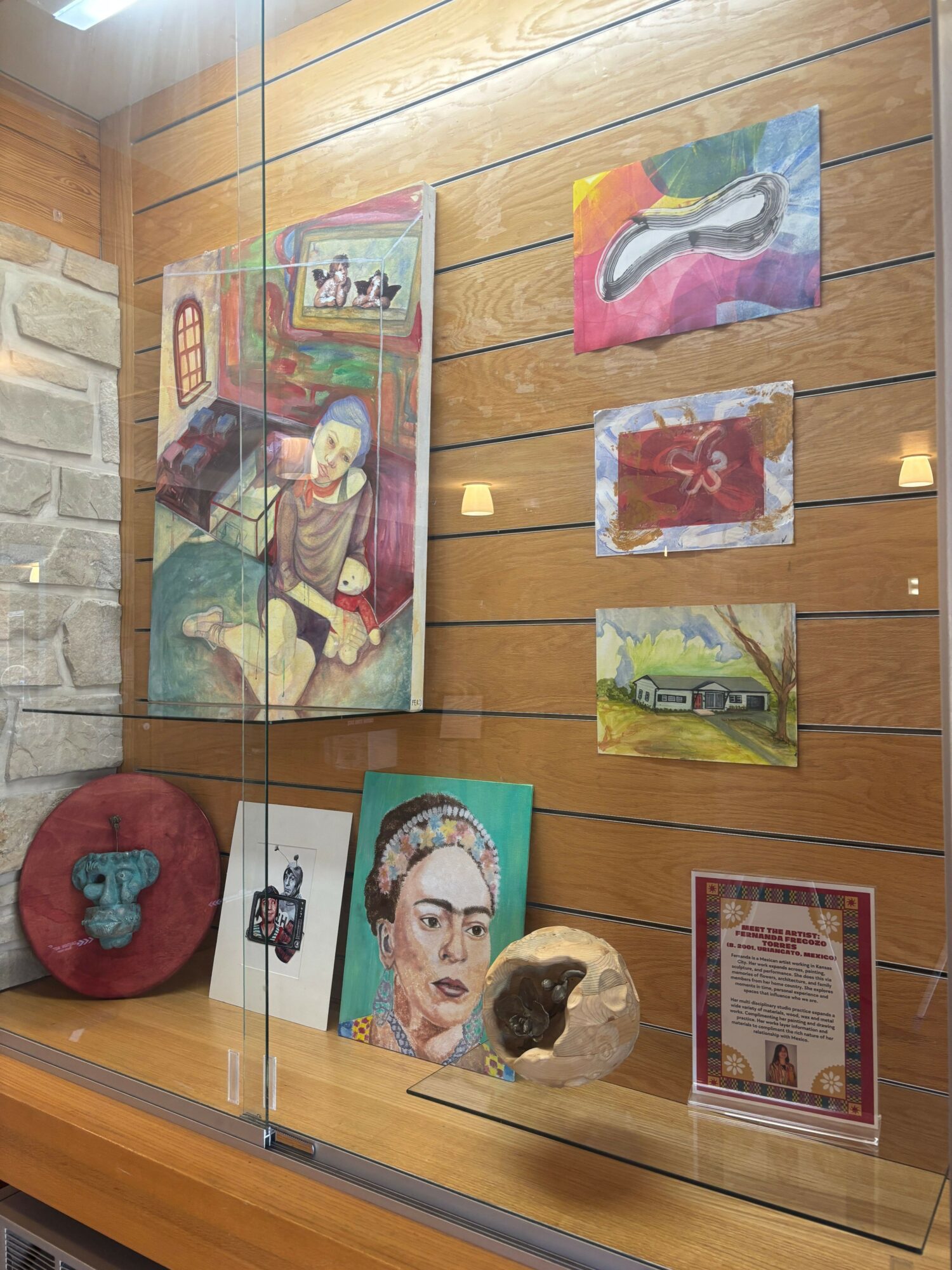 Display of colorful artwork, a portrait, and a small sculpture on a wooden wall and glass shelf.