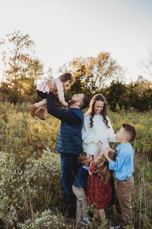 Family outdoors with a man lifting a girl, woman and two boys standing nearby, trees and sky in background.