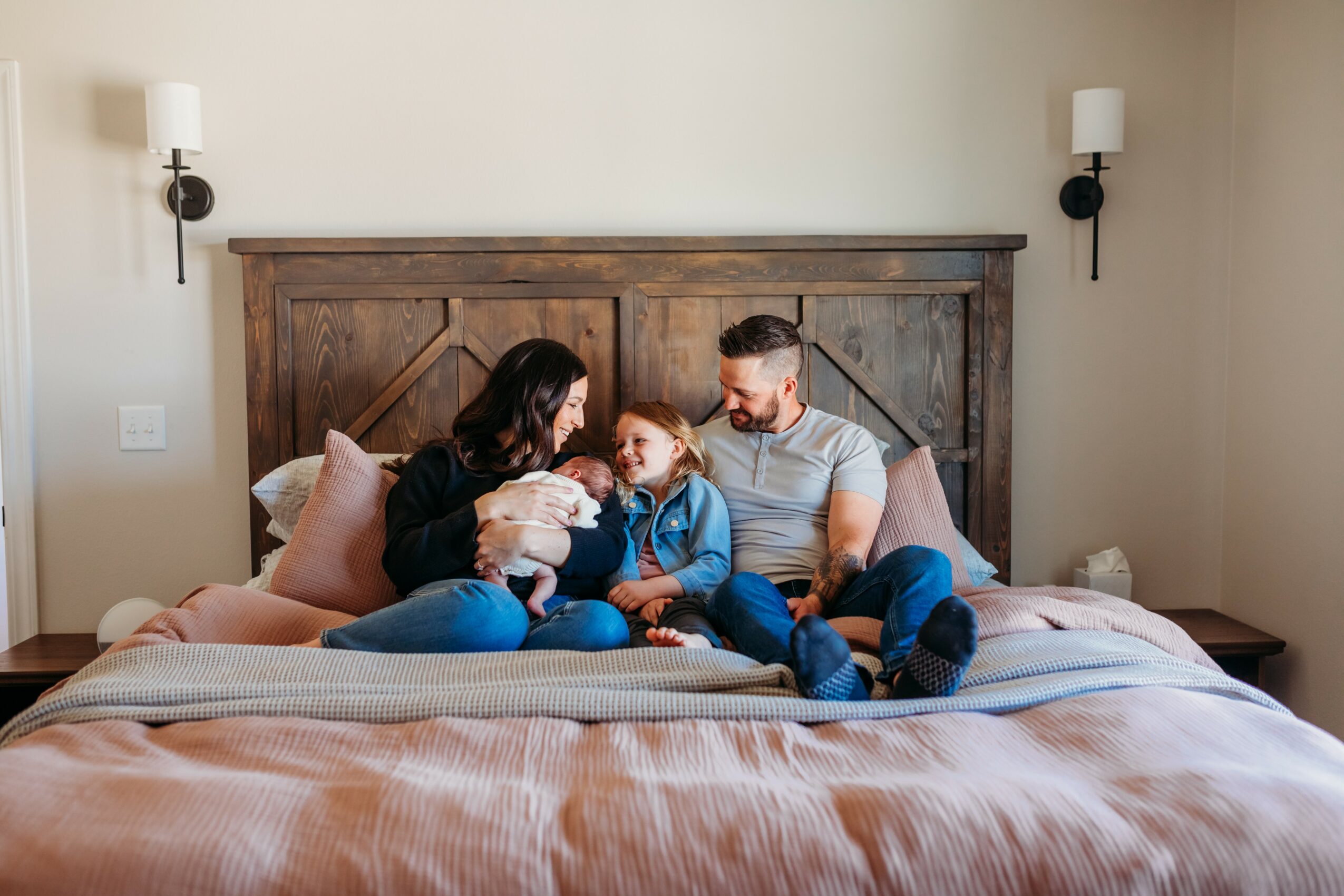 Family of four sitting on bed, holding a small dog, in a bedroom with wooden headboard and wall-mounted lamps.