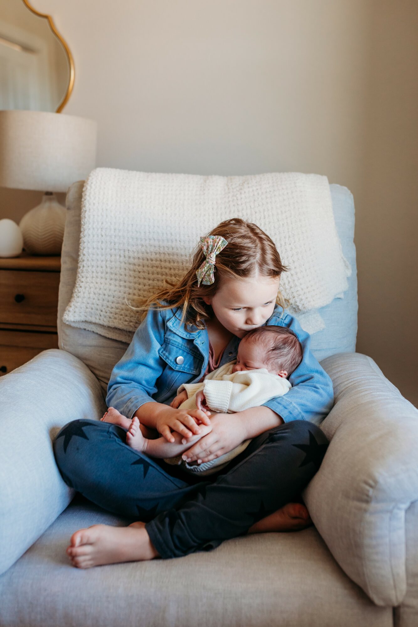 Young girl sitting on a chair holding a baby, both looking at each other, in a cozy room with a lamp and side table.