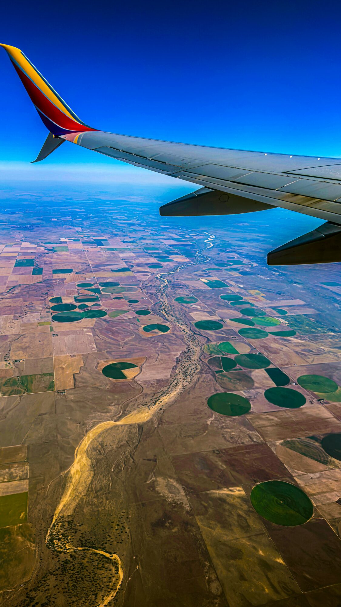 Airplane wing with colorful tail flying over patchwork farmland and a winding river, under a clear blue sky.