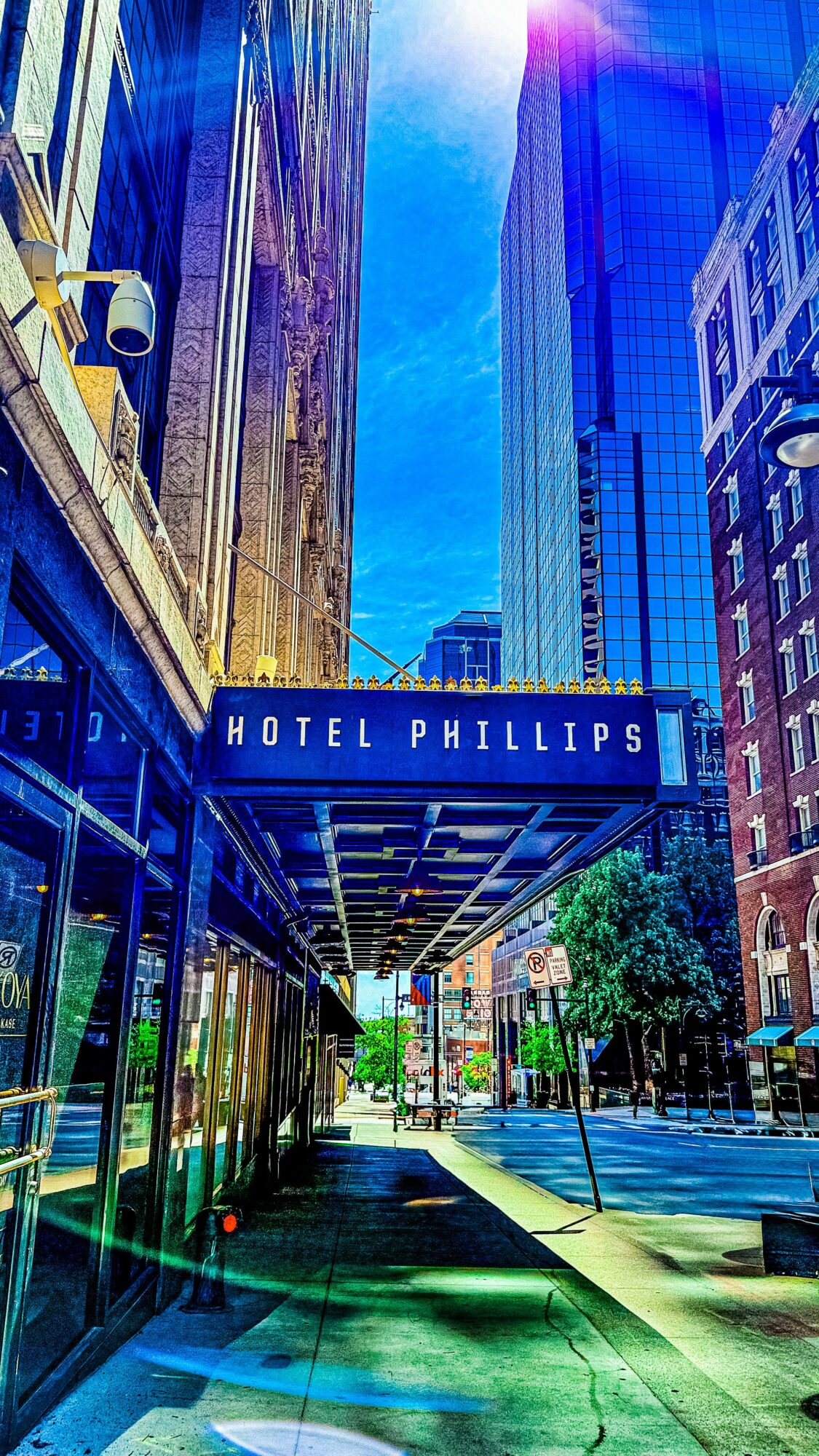 City street with tall buildings, hotel Phillips sign, trees, and a sidewalk, under a blue sky.