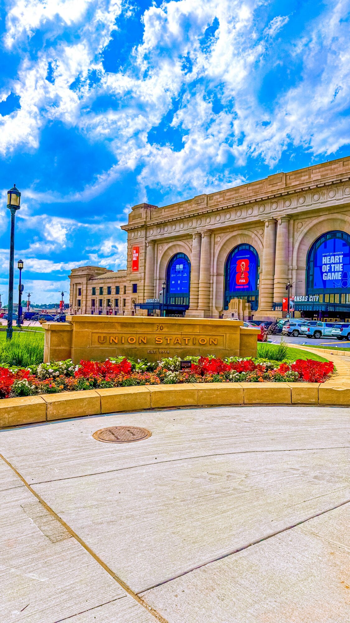 Building with large arched windows, blue banners, and a flower bed in front, under a partly cloudy sky.