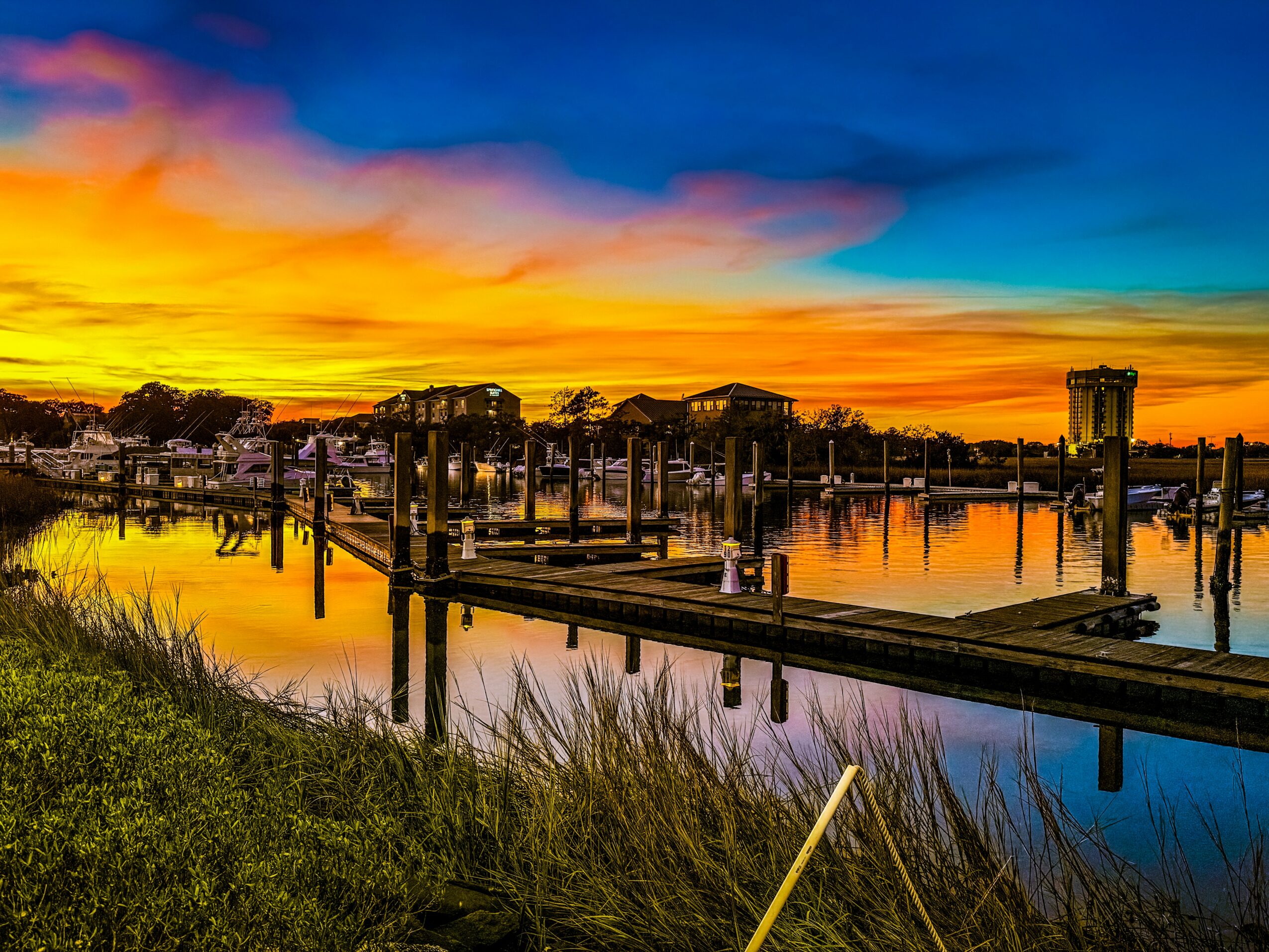 Sunset over a marina with boats, docks, and calm water reflecting the colorful sky, with grassy foreground.