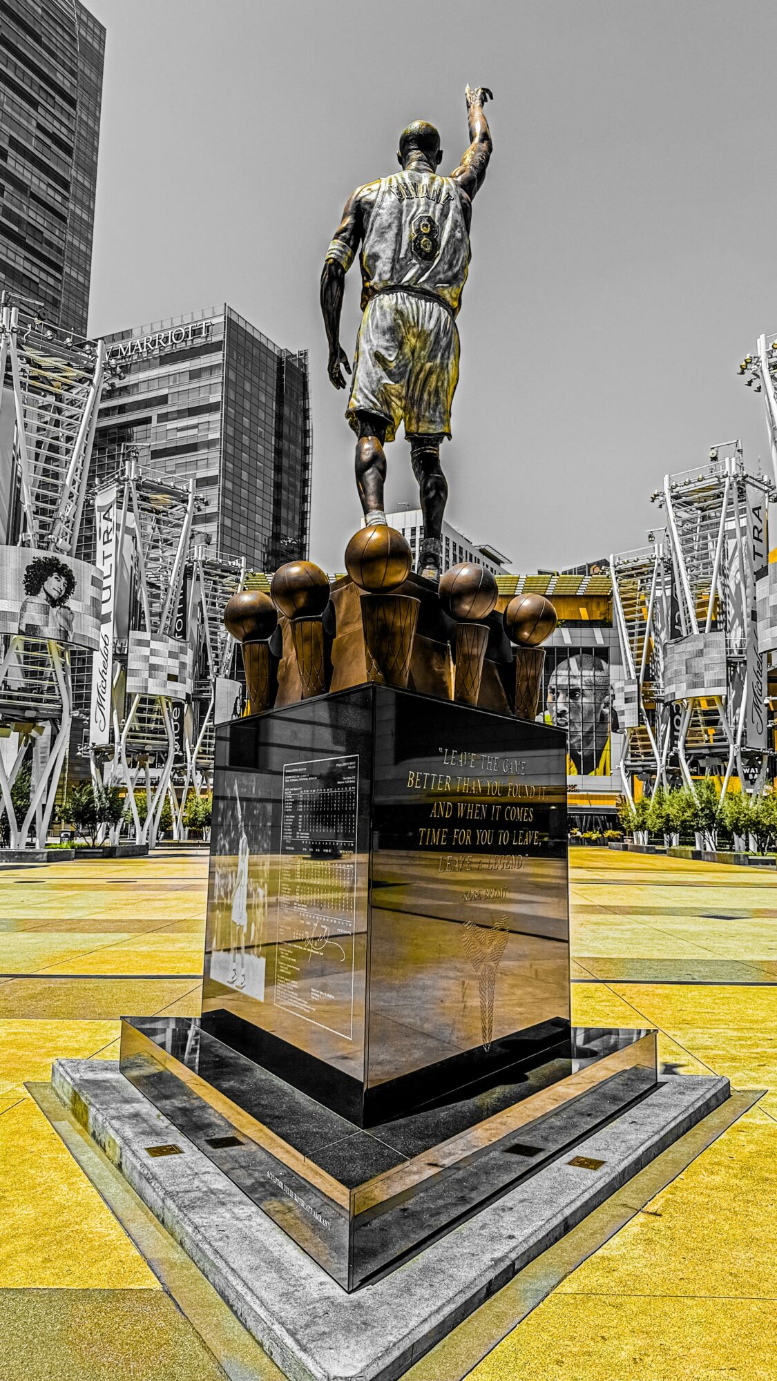 Statue of a person with one arm raised, standing on a pedestal in an urban park with modern buildings in the background.