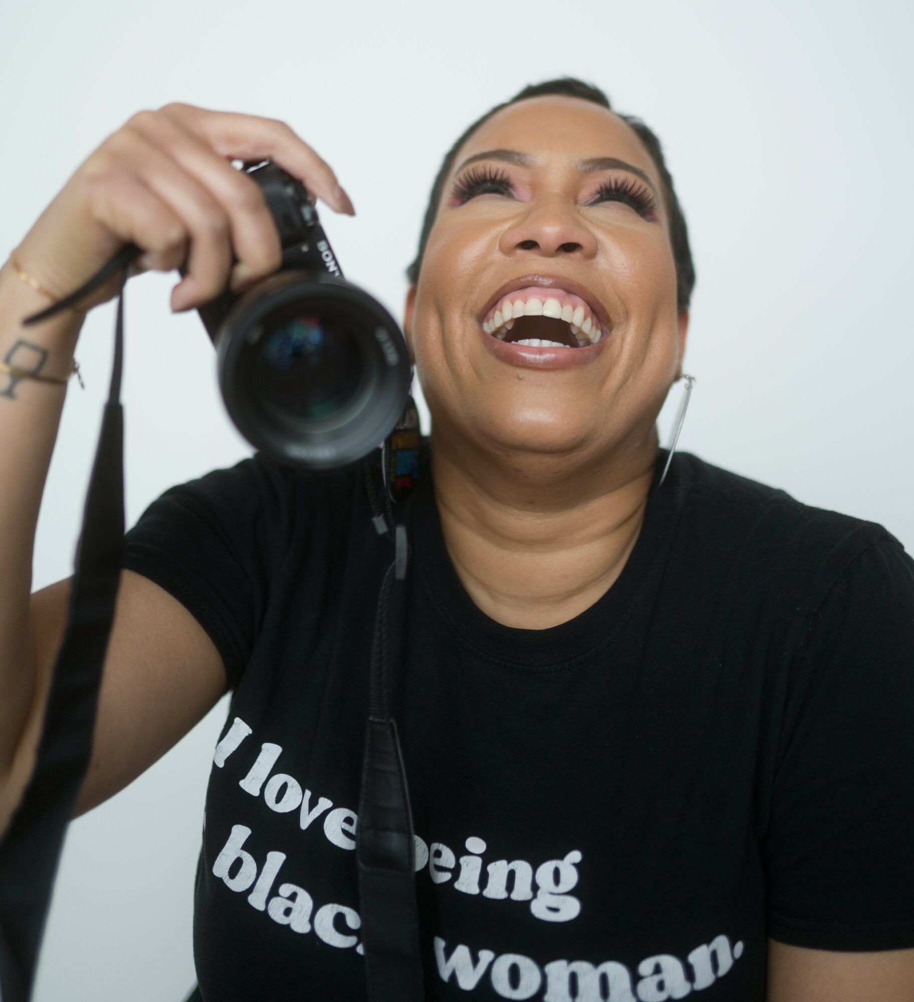 Woman smiling and holding a camera, wearing a black shirt with white text, against a plain background.
