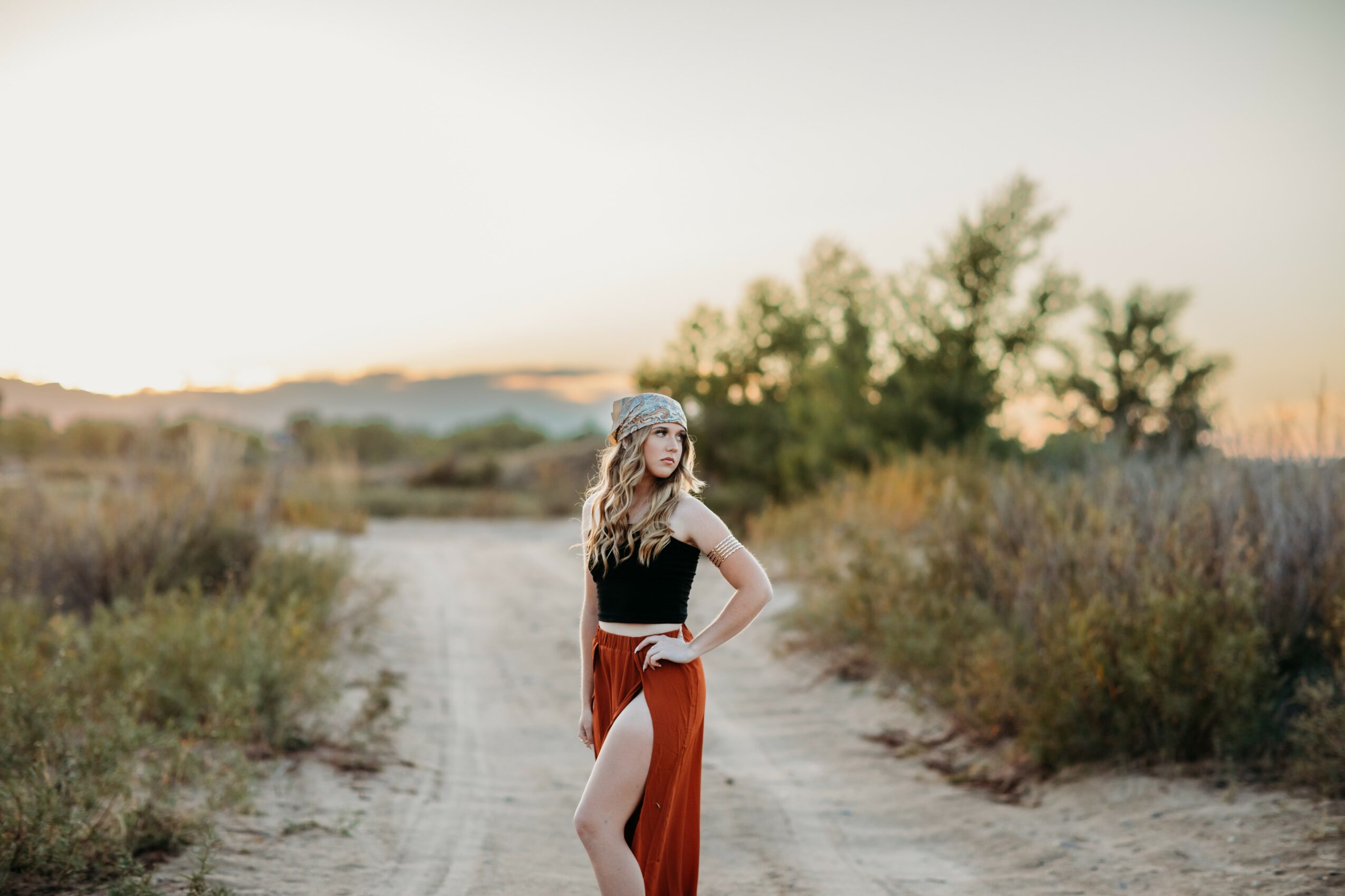Woman standing on a dirt path in a natural landscape during sunset, wearing a black top and red skirt with a slit.