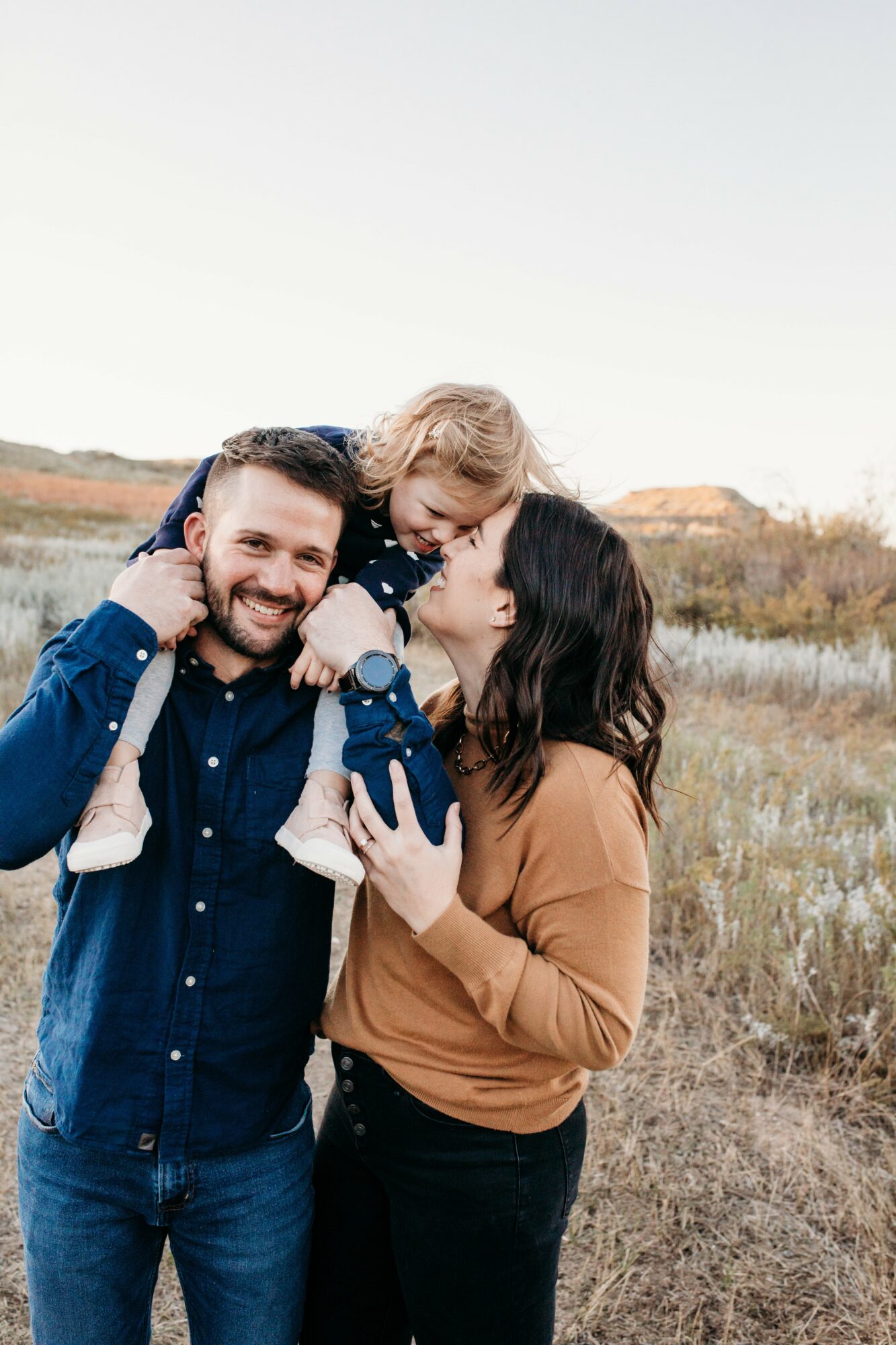 Family of three outdoors, woman holding child, man smiling, grassy field background, clear sky.
