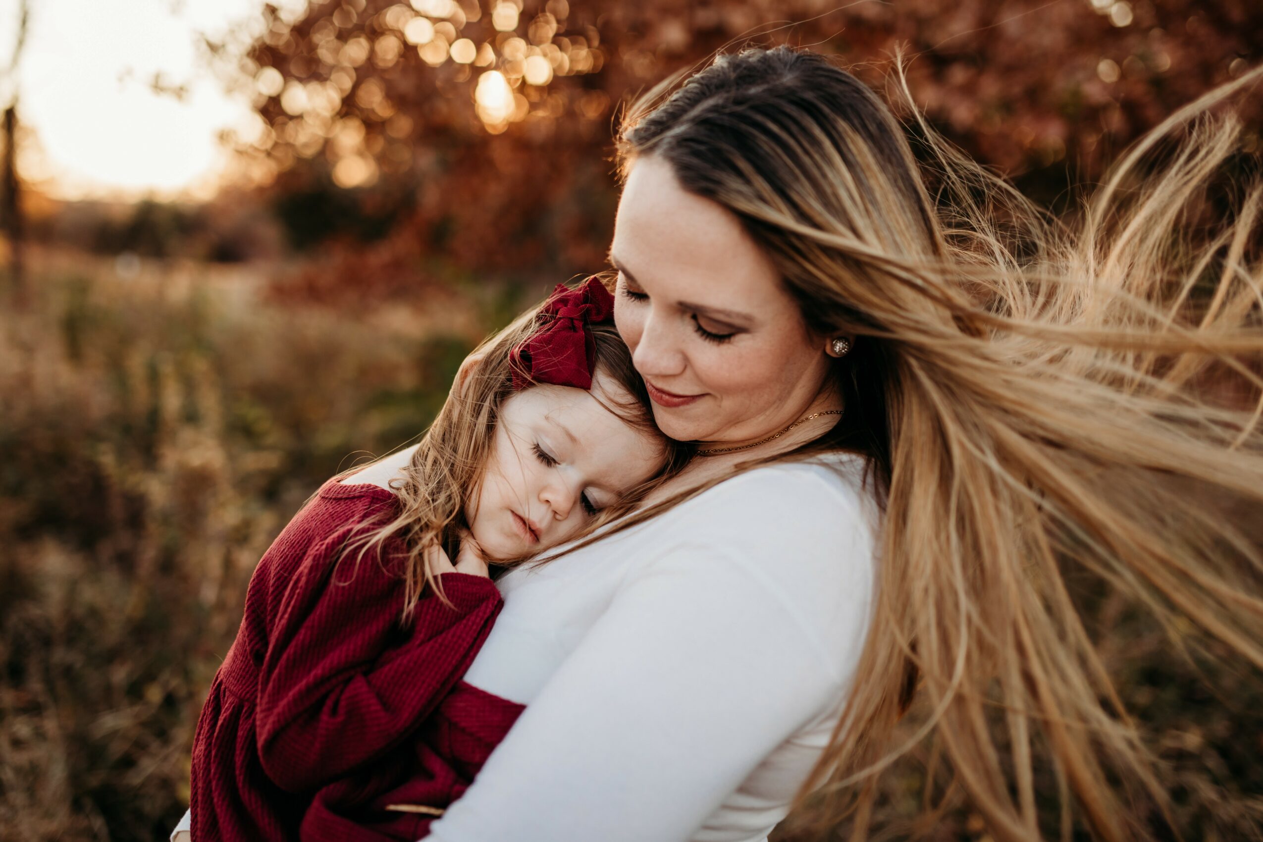 Woman holding a sleeping child outdoors during autumn, with trees and warm sunlight in the background.