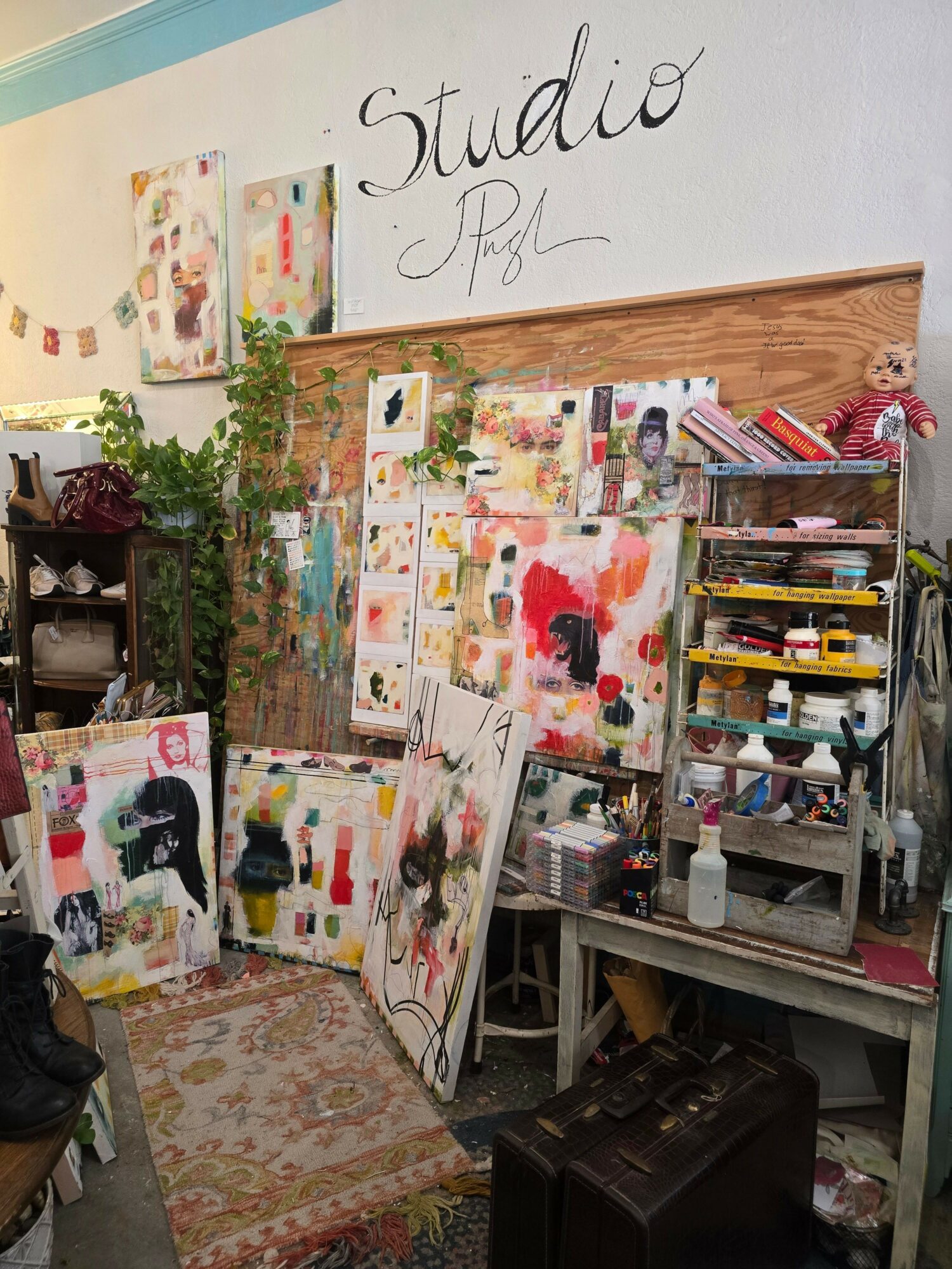 Art studio with colorful paintings, supplies, and a plant on a table, against a wooden wall with handwritten sign.