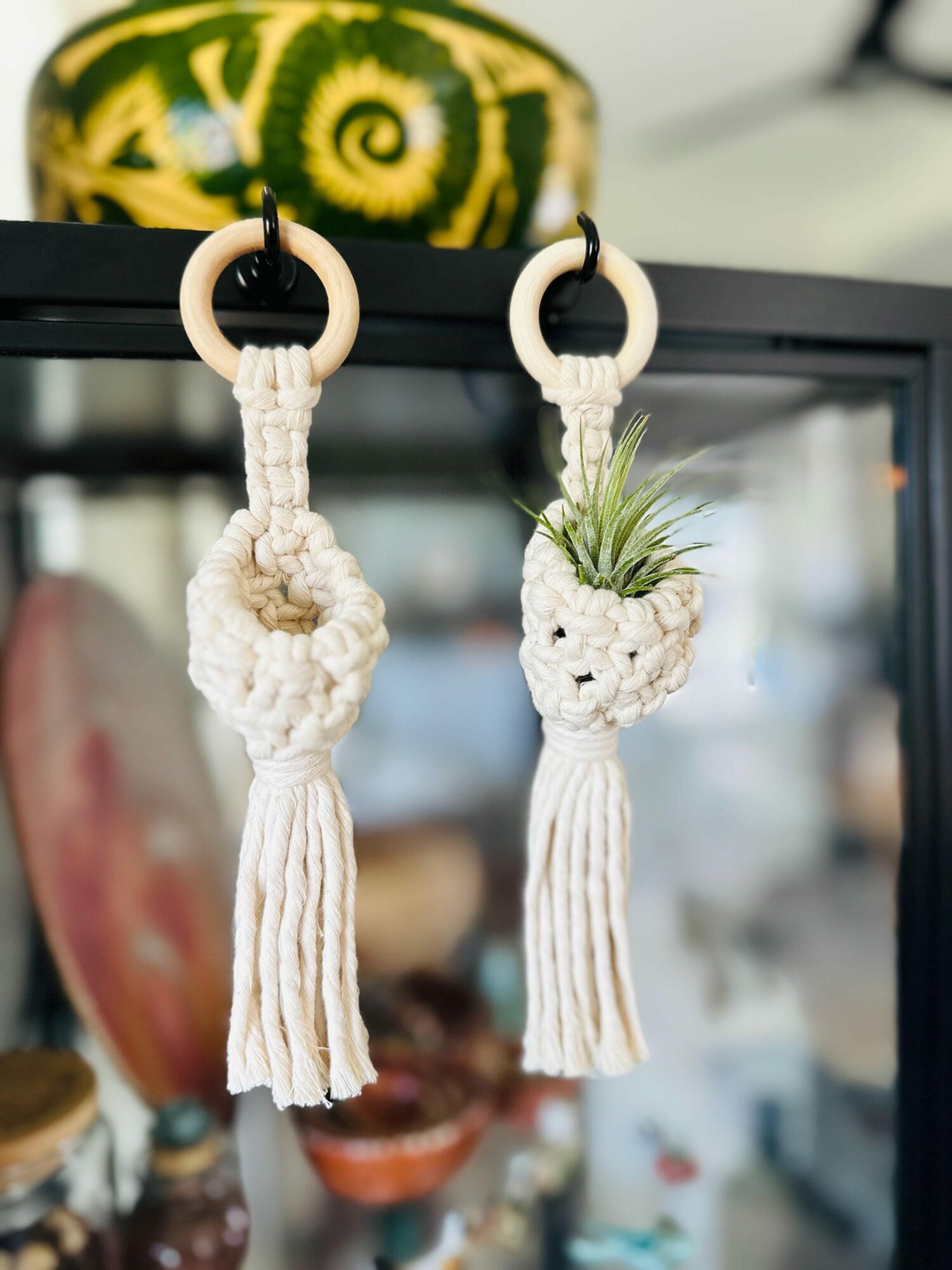 Two macrame pineapple keychains hanging from black rings, with a blurred background including a watermelon and other objects.