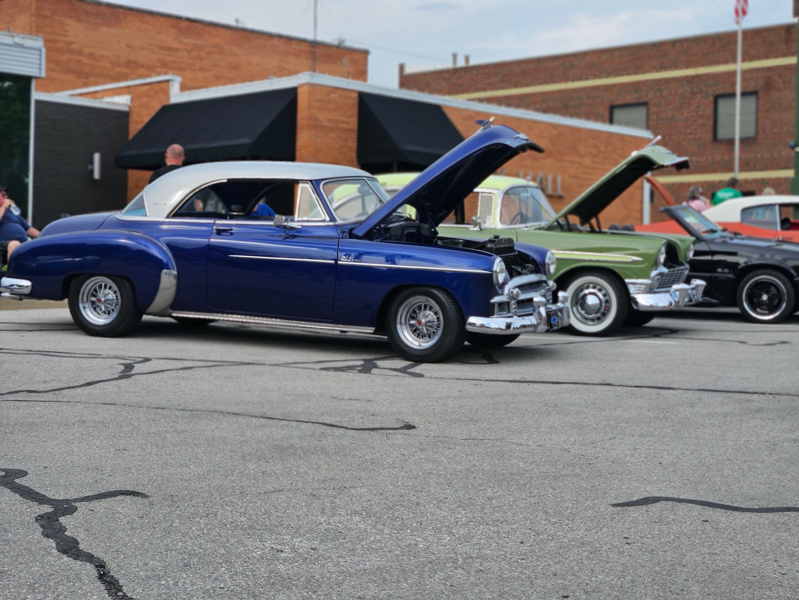 Blue vintage car with open hood parked on street, other classic cars and buildings in background.
