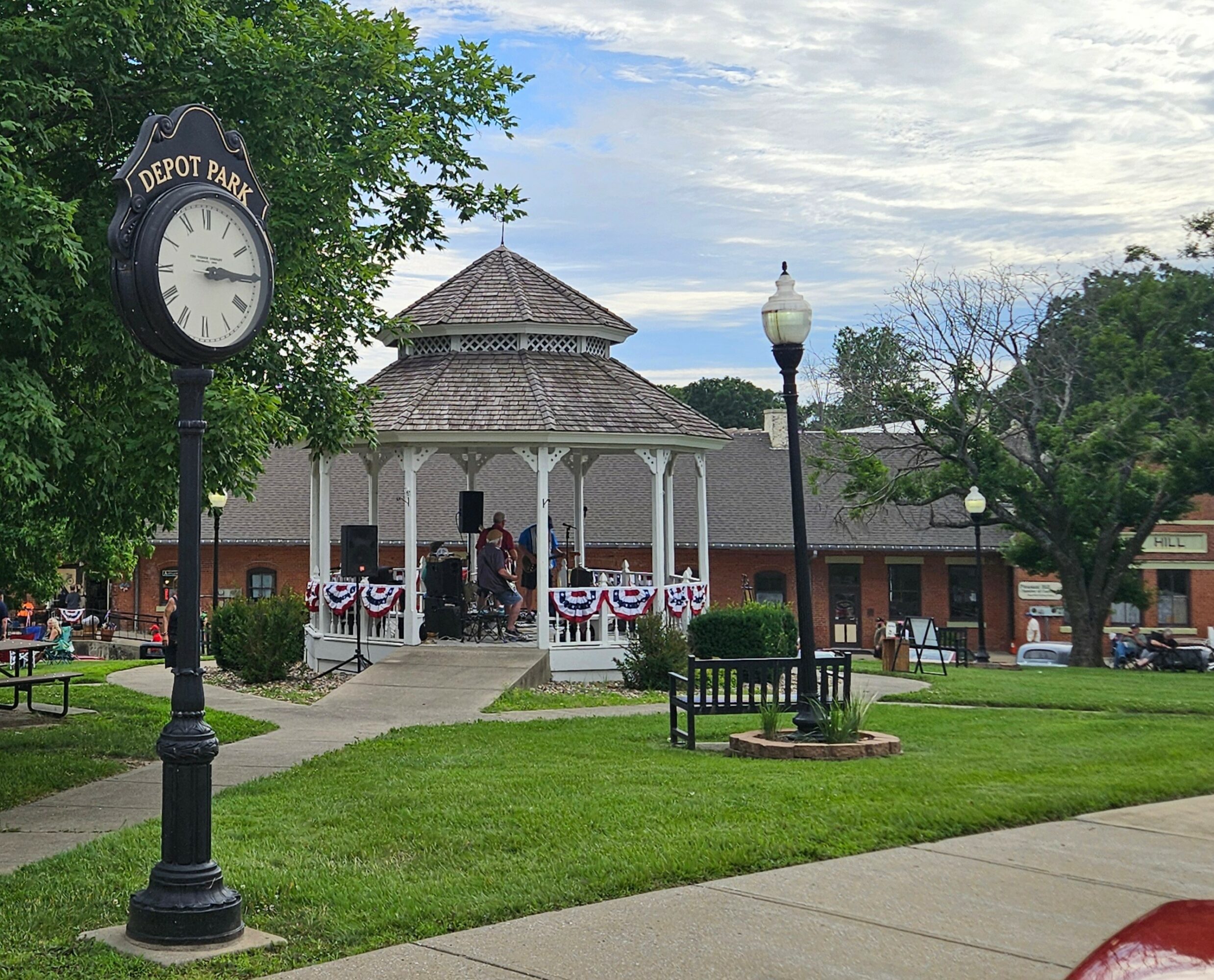 Park with clock, gazebo, benches, and trees, with a band playing on the gazebo, and a partly cloudy sky.