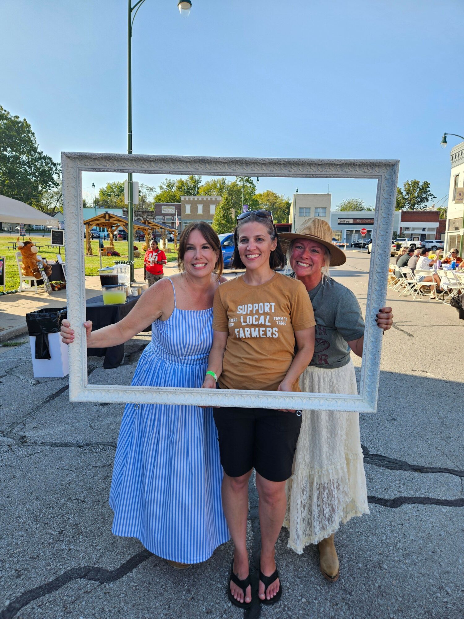 Three people posing with a large empty picture frame outdoors, with a fair or festival in the background.