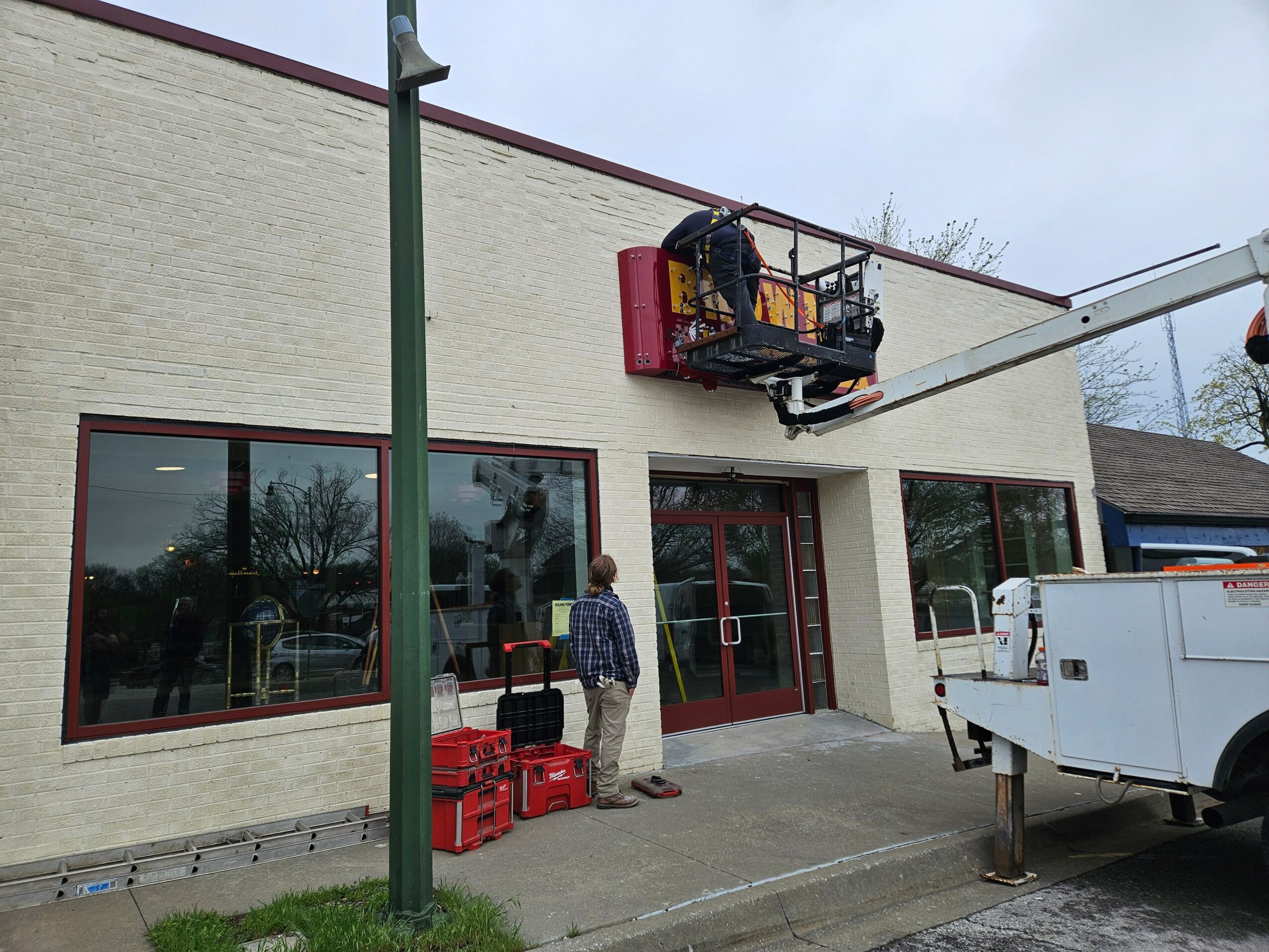 Person on lift working on building exterior wall, with crates on ground nearby, in front of large windows and door.
