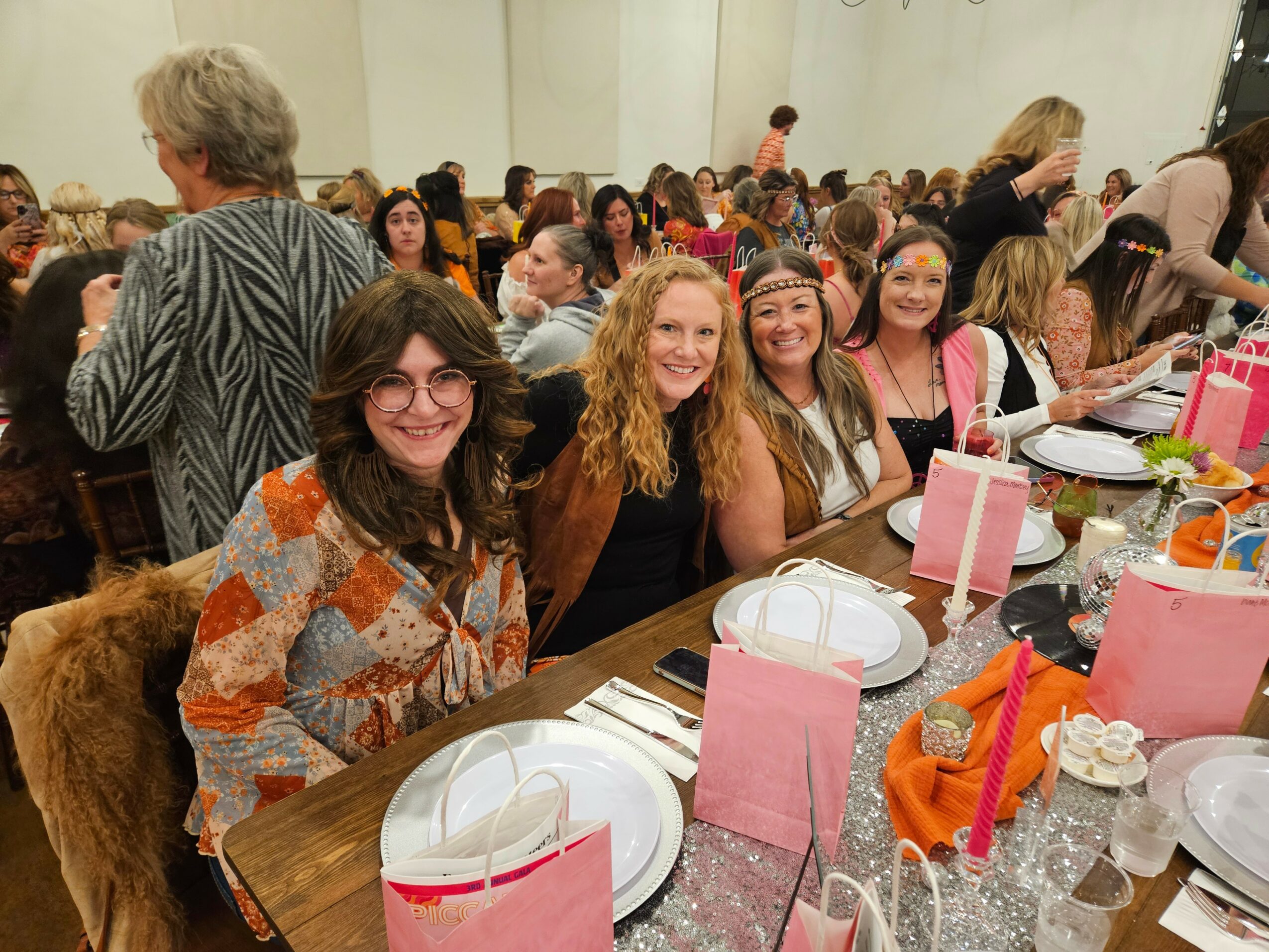 Group of smiling women sitting at a table during a social event, with pink gift bags and tableware visible.