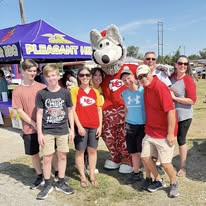 Group of children and adults with a mascot wolf at an outdoor event, standing in front of a purple tent, smiling.