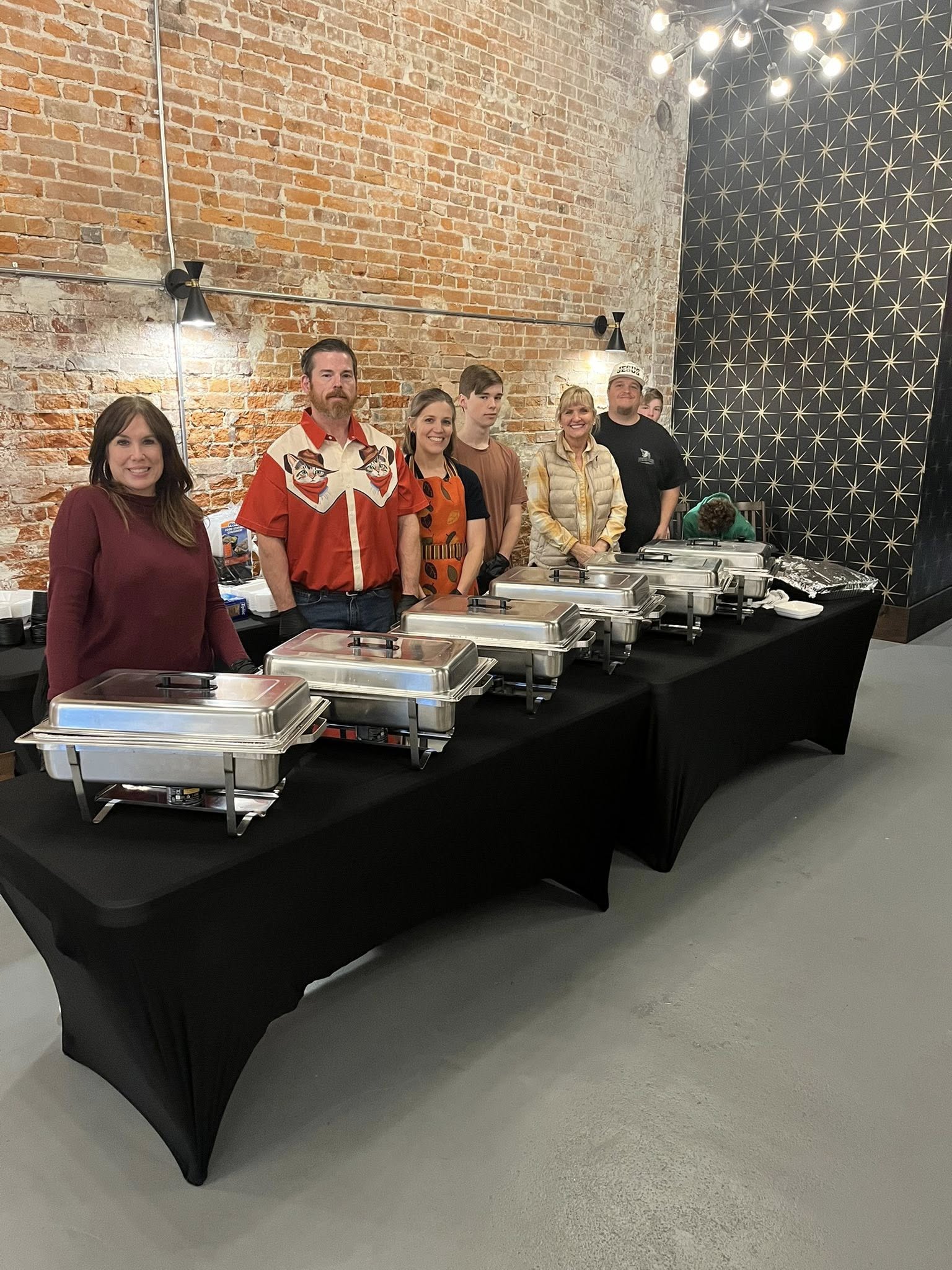Six people stand behind a buffet table with chafing dishes in a brick-walled room, some smiling at the camera.